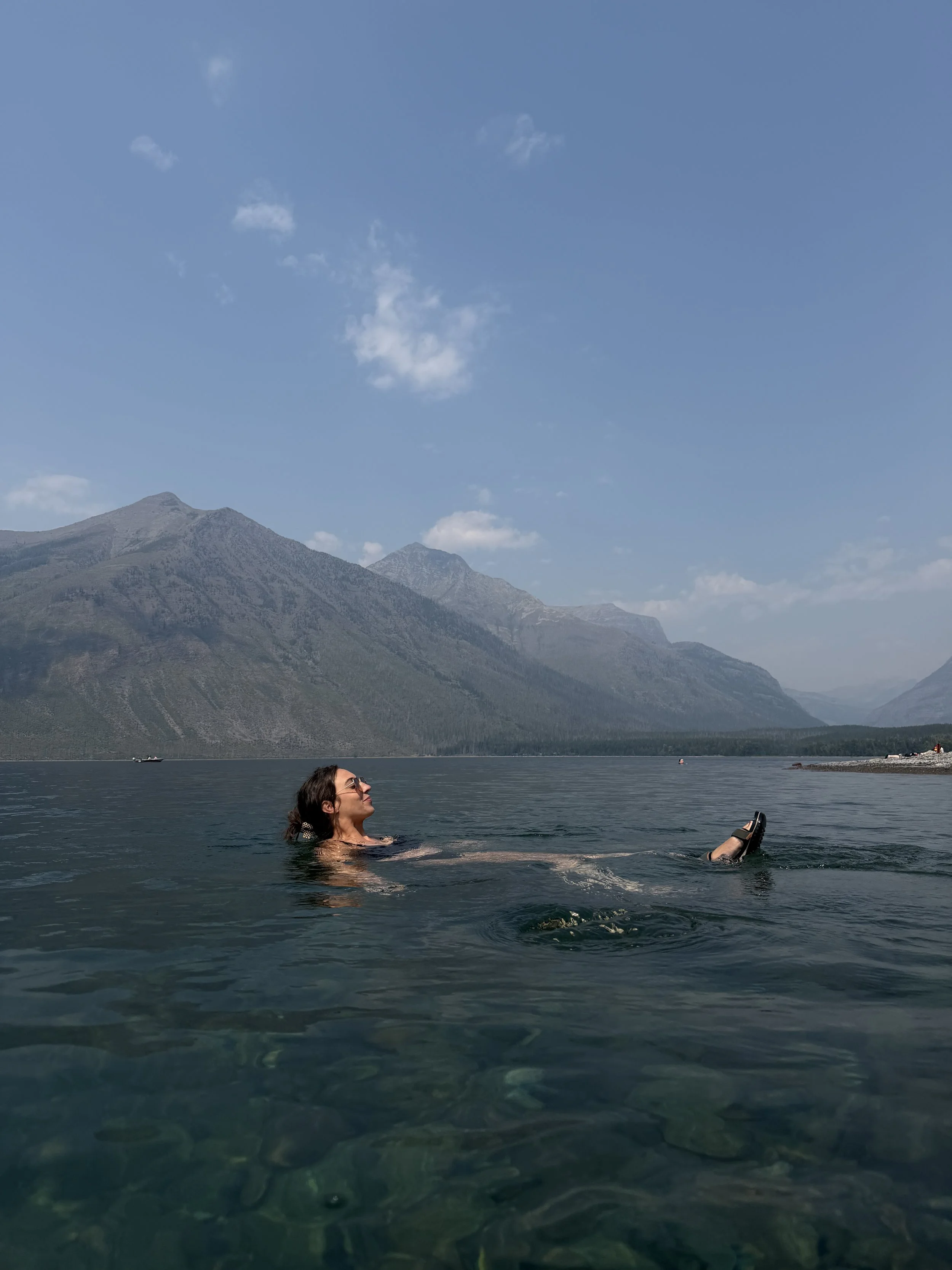 A woman with dark hair, wearing sunglasses and a ring, is relaxing in a body of water, with mountains and a blue sky with some clouds in the background.