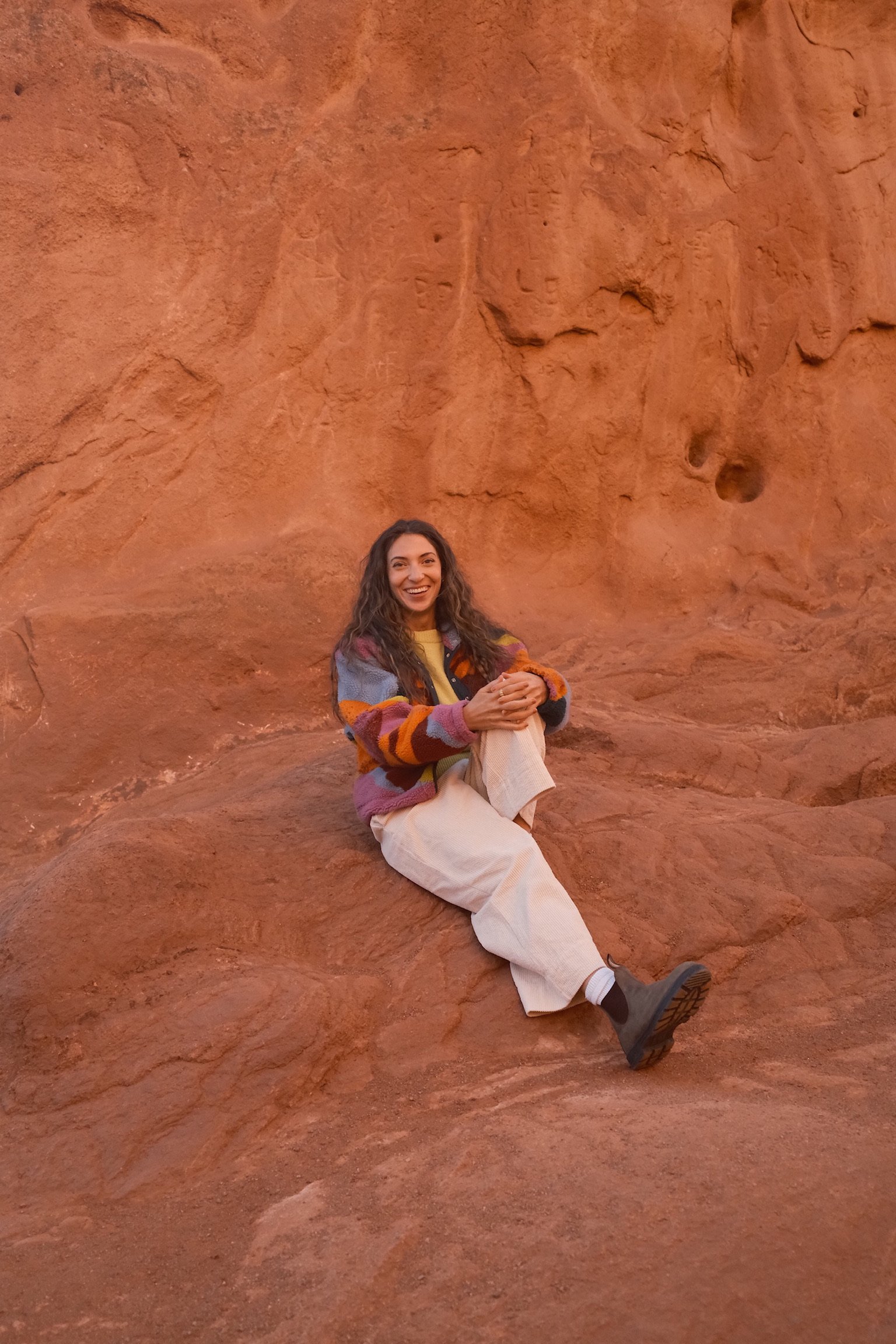 A smiling woman with long, curly hair sitting on reddish-brown rock formations, wearing a colorful patchwork sweater, white pants, and black boots.