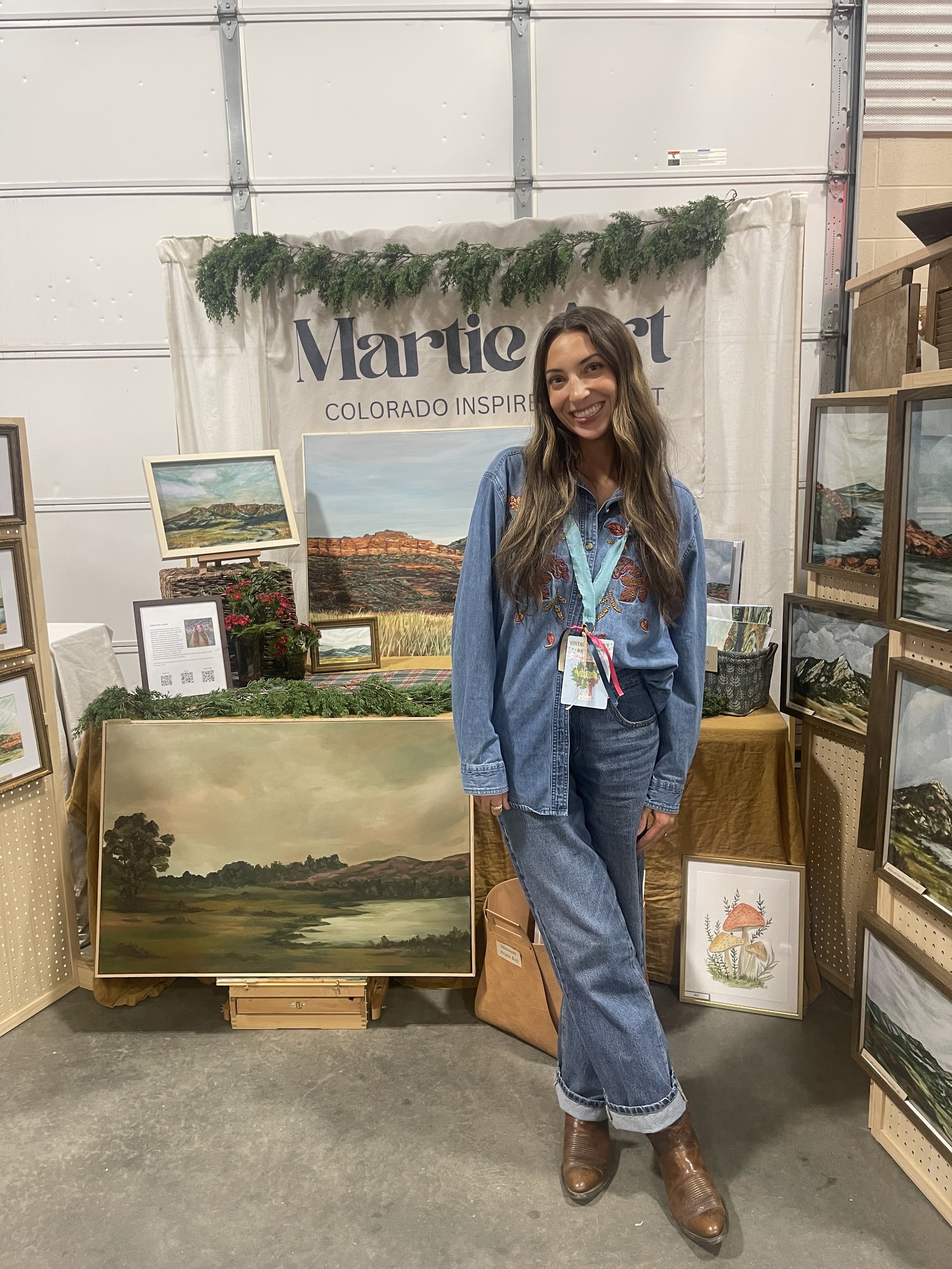 A woman standing in front of an art display at a booth with paintings of landscapes and nature scenes. She is smiling, wearing a denim shirt and jeans, with a lanyard around her neck.
