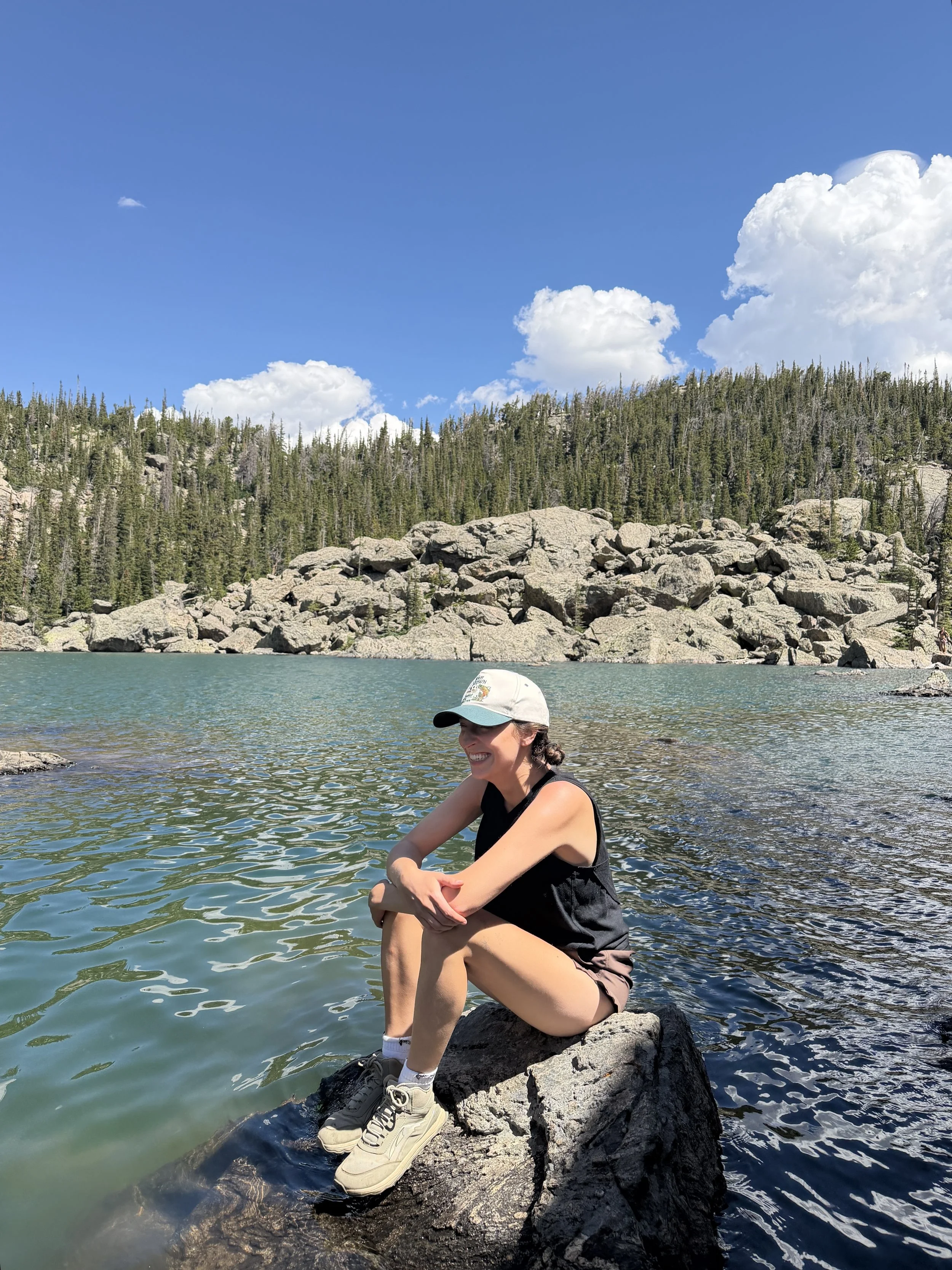 A woman in a black tank top, light-colored shorts, beige sneakers, and a white cap sits on a rock by a lake with a mountain and forest background under a blue sky with clouds.