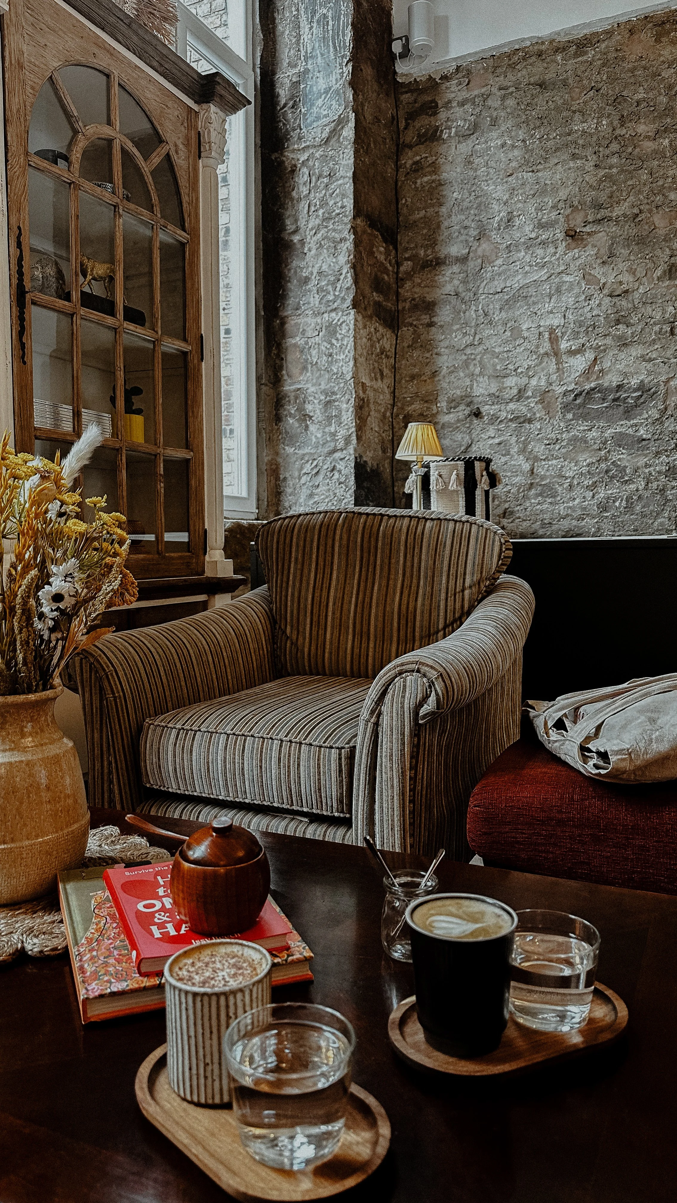 Cozy coffee shop scene with striped armchair, wooden table with drinks, flowers in a vase, books, and rustic stone wall.