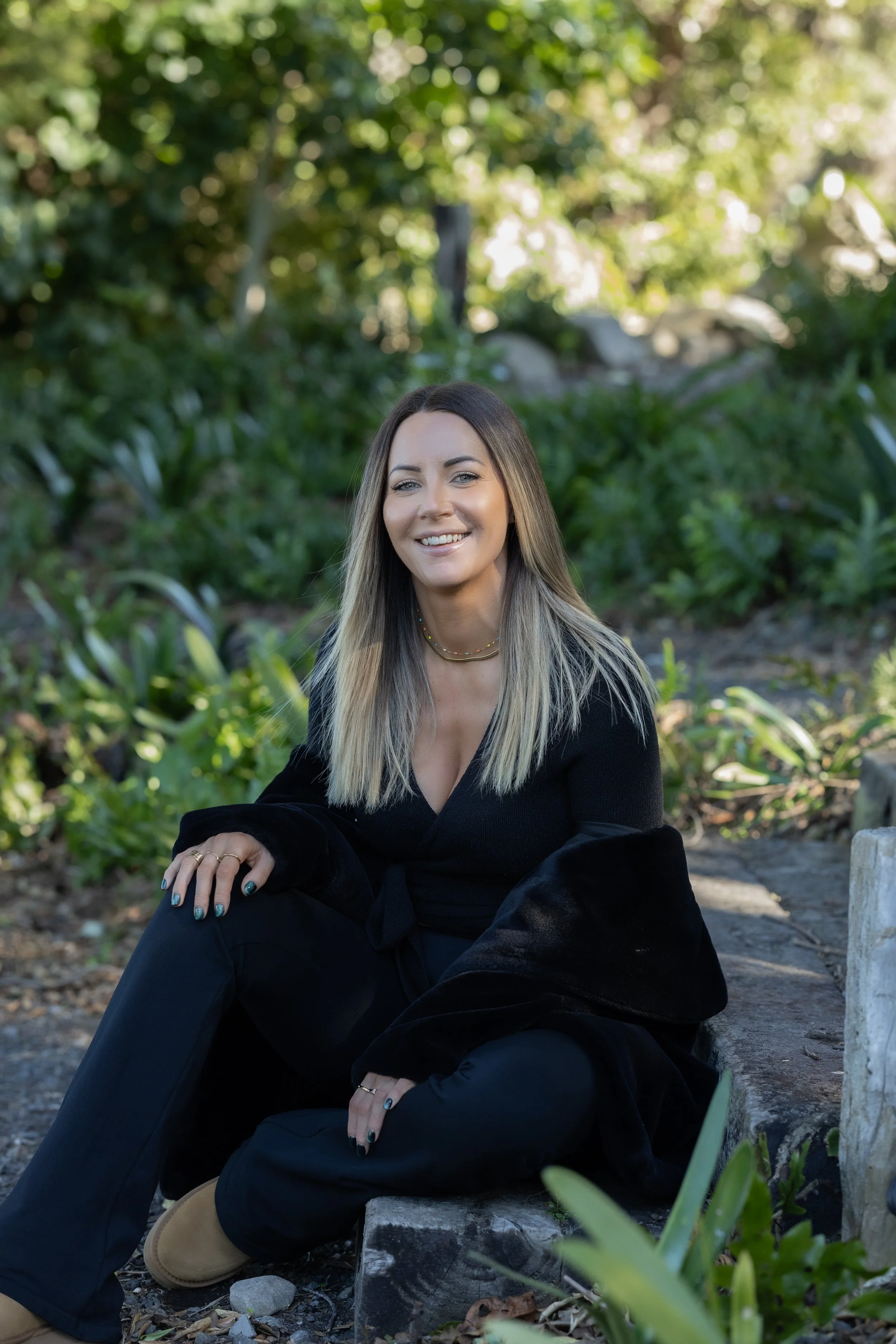 A woman with long, blonde hair and light makeup sitting outdoors on a rock, smiling at the camera. She is wearing a black outfit with a layered necklace, surrounded by greenery and trees.