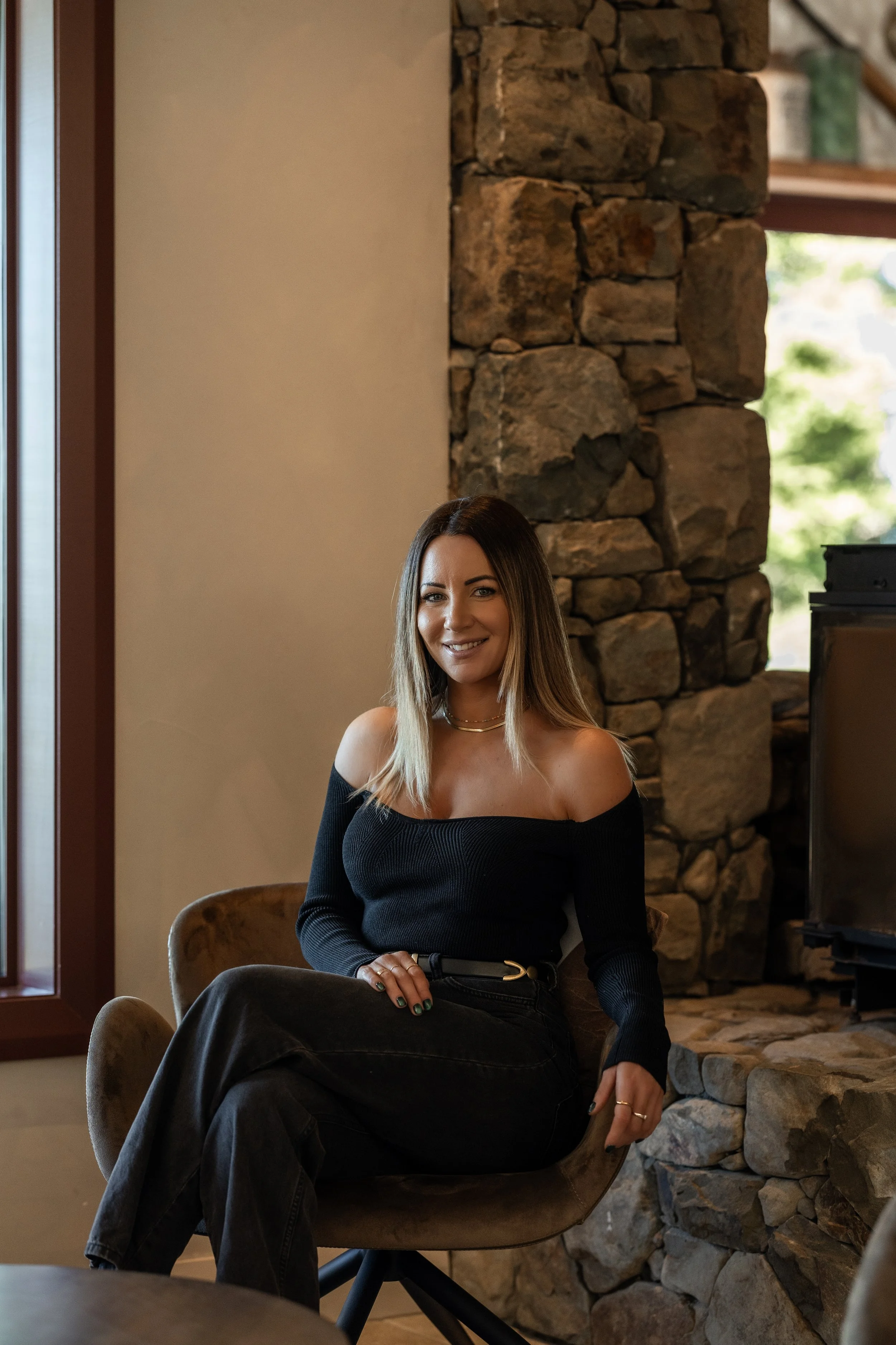A woman sitting on a beige chair near a stone fireplace in a cozy indoor setting.