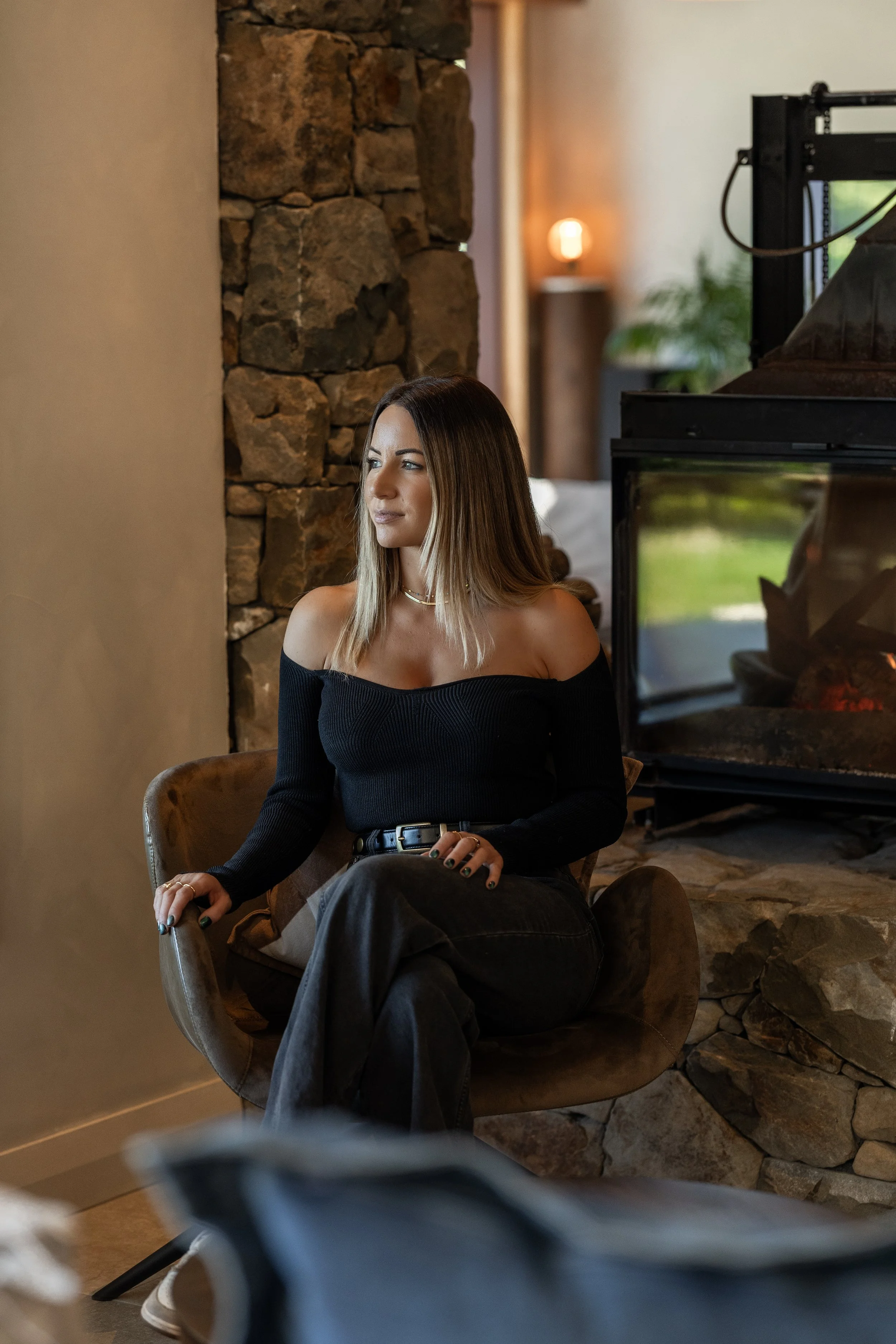 A young woman with shoulder-length blonde hair, wearing a black off-the-shoulder sweater and black pants, sits in a cozy living room by a stone fireplace, looking to the side.