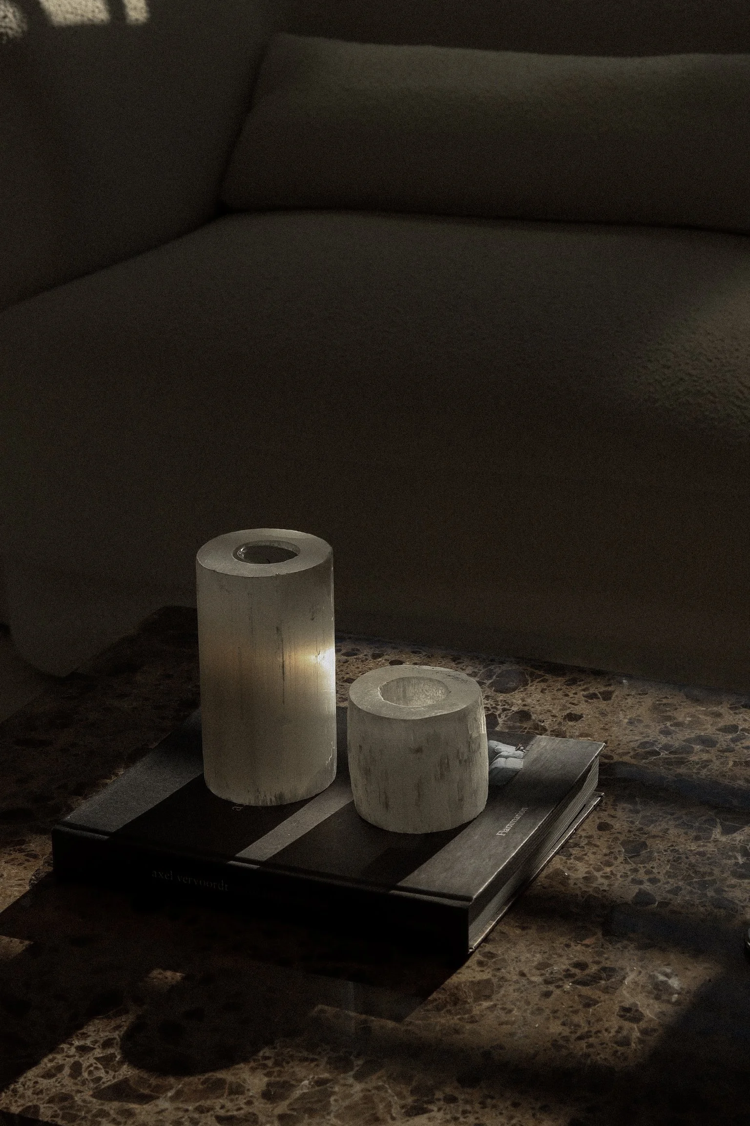 Two white candles on top of a closed book, on a marble surface with a sofa in the background.