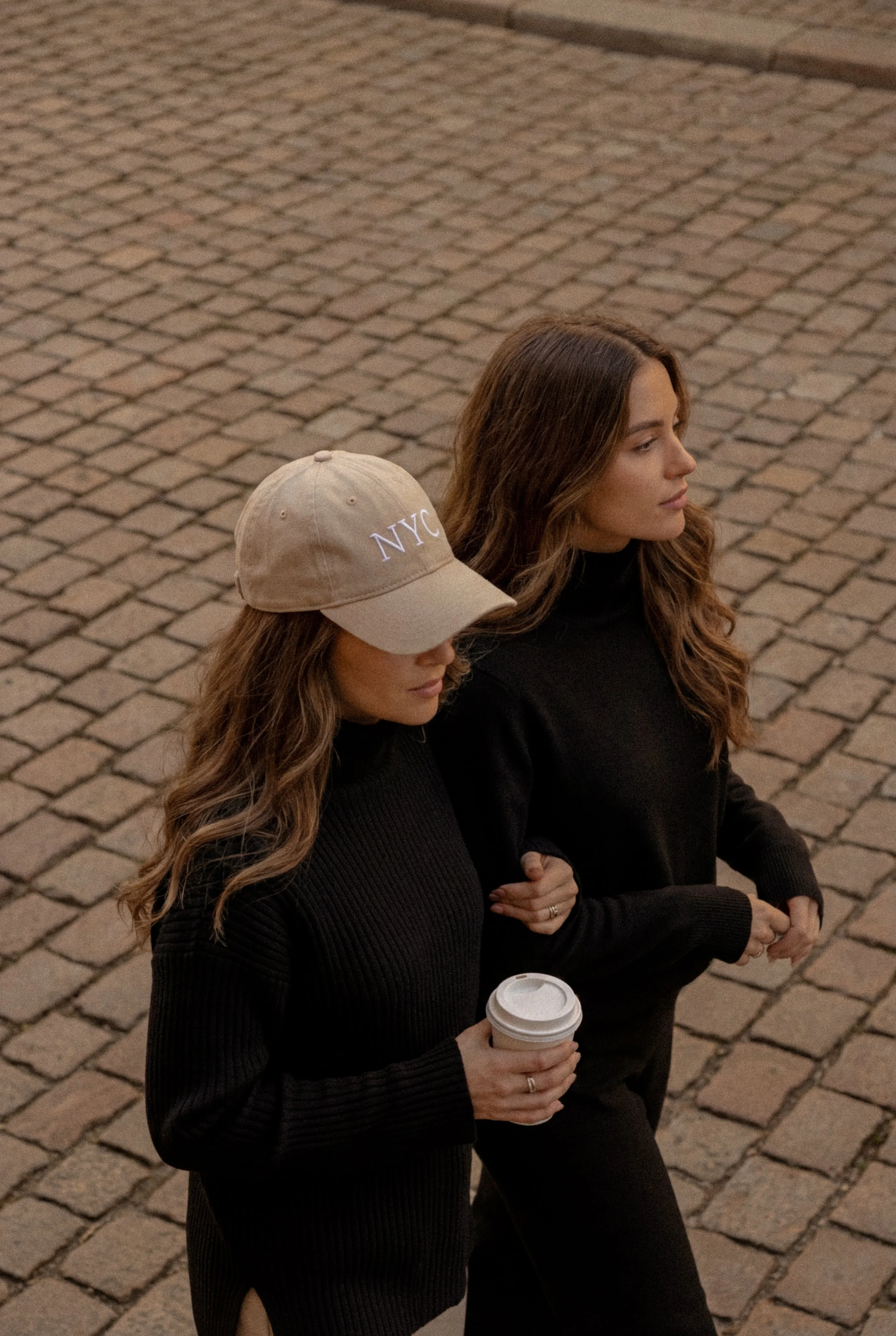 Two women walking on a cobblestone street, one wearing a beige NYC cap and holding a coffee cup, the other dressed in a black sweater. The woman wearing the cap has long, wavy hair, and the other has long, wavy brown hair.