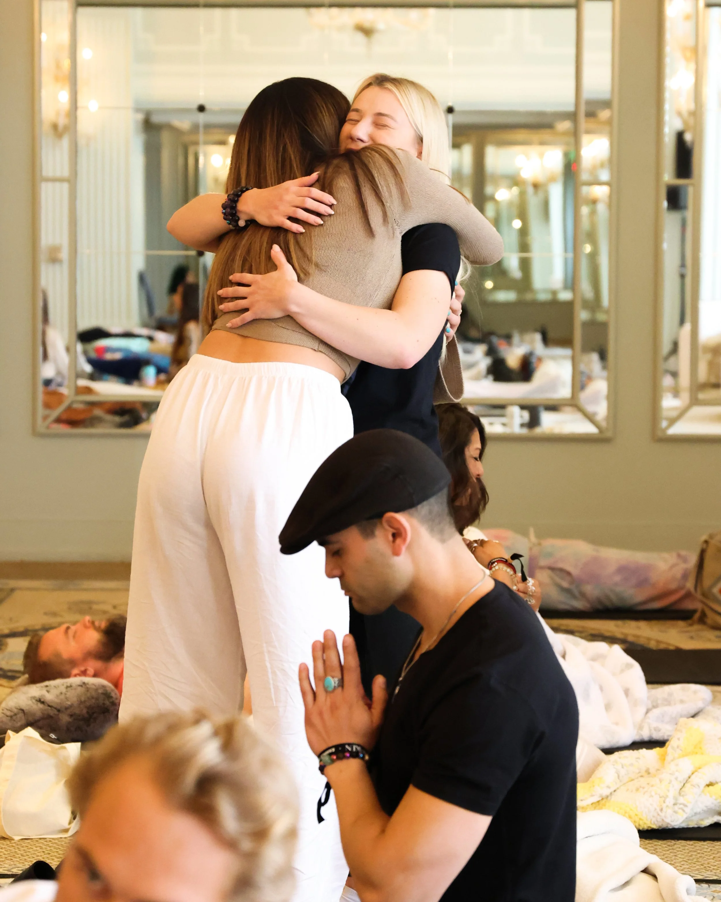 People hugging and praying in a spiritual gathering, some lying on the floor, with a mirror reflecting the room's interior.