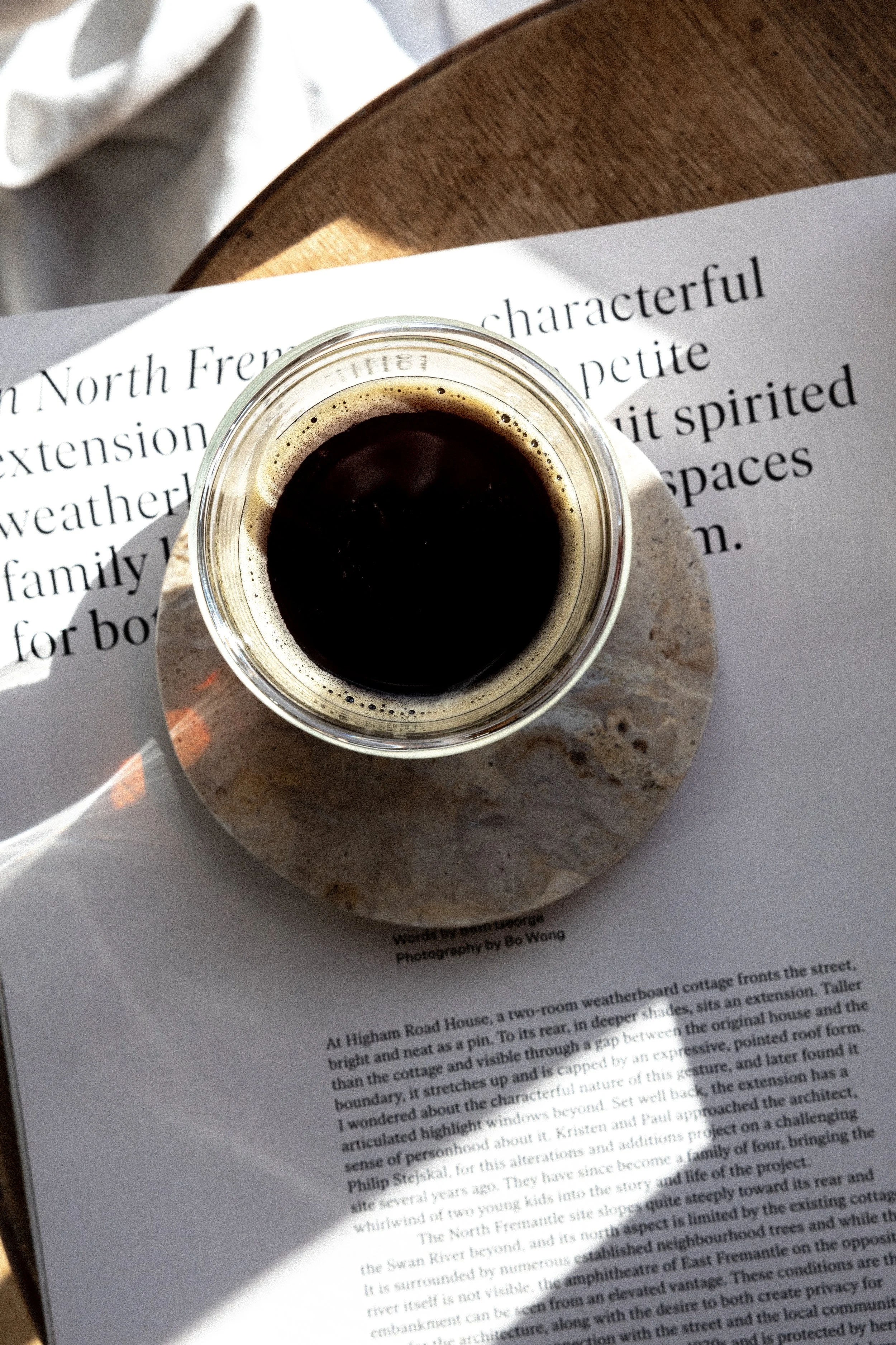 Top view of a glass of black coffee on a marble coaster, placed on an open book with text, on a wooden surface.