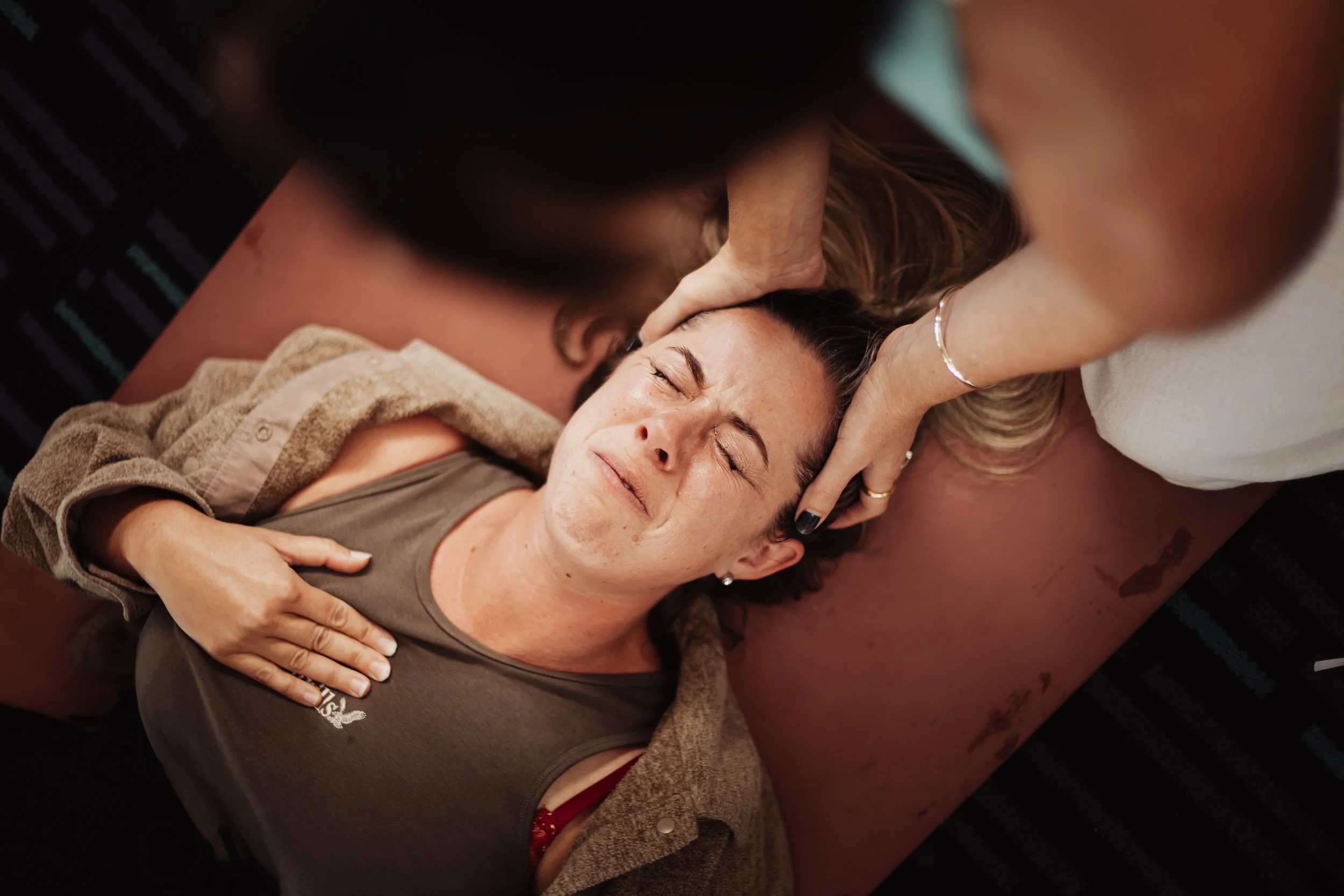 Woman lying on a massage table with her eyes closed, experiencing discomfort, while a person performs a facial or massage treatment on her forehead.