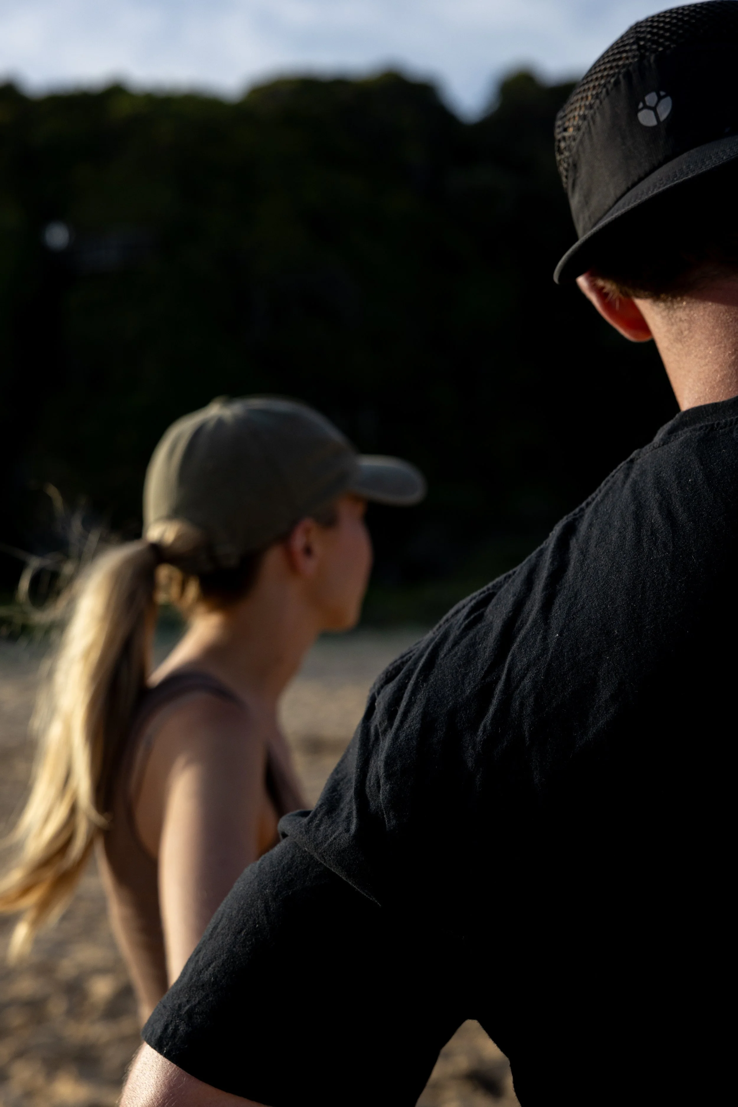 Back view of a man and a woman standing outdoors, both wearing caps. The woman's hair is in a ponytail, and they are on a sandy area with a dark, blurry background.