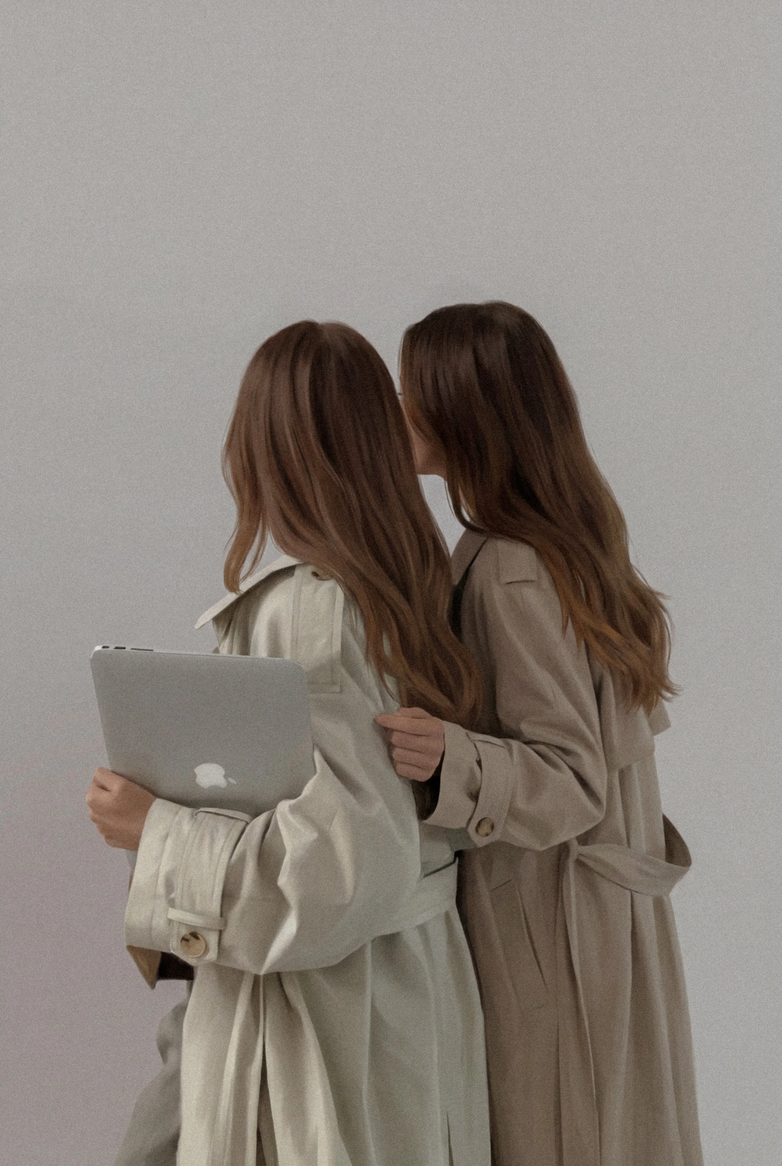 Two women with long hair, wearing beige trench coats, are close together, possibly kissing or whispering, with one woman holding a silver laptop.