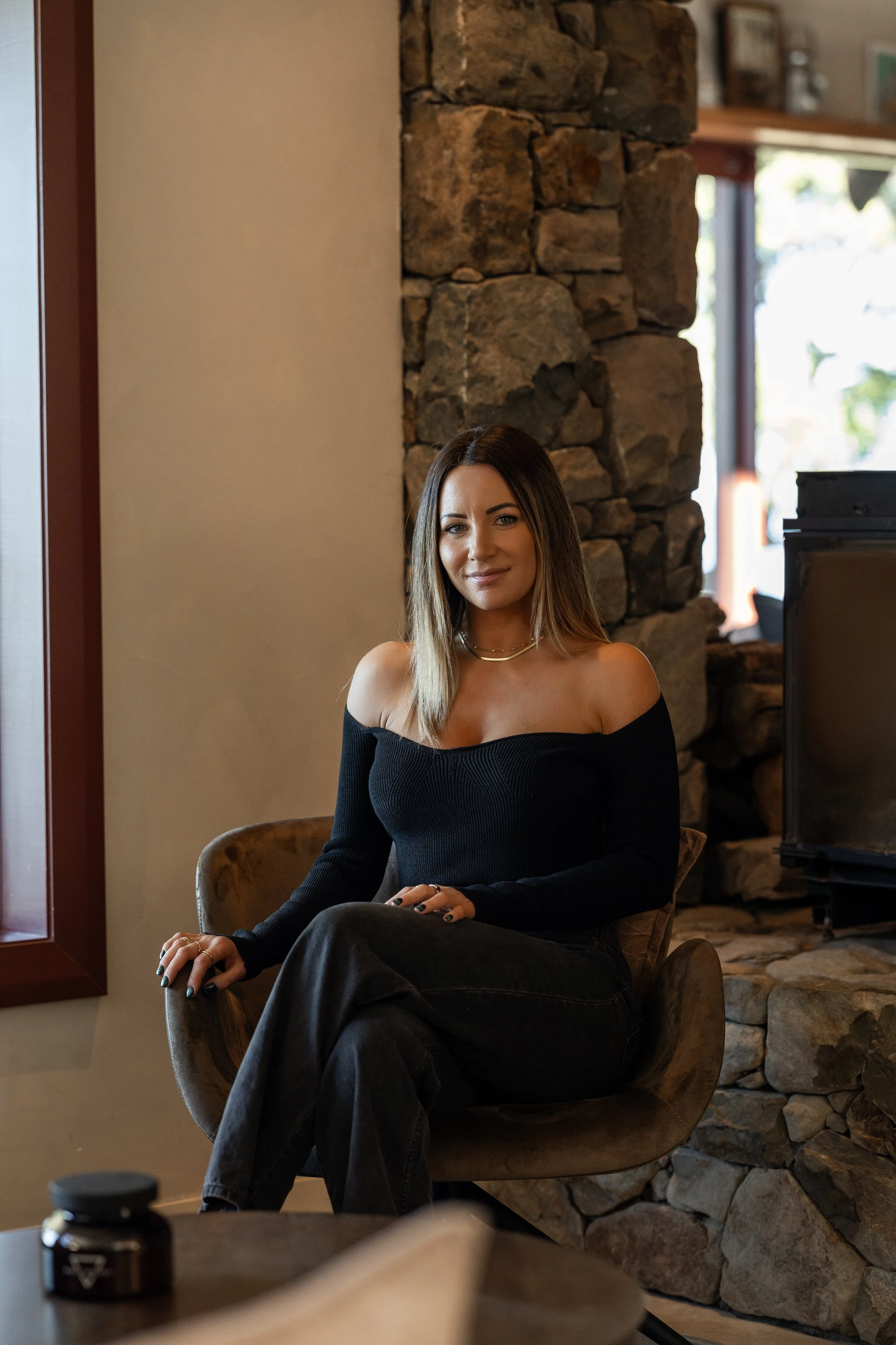 A woman with shoulder-length hair, wearing an off-the-shoulder black top and black pants, sits on a beige chair inside a cozy room with a stone fireplace and large window.