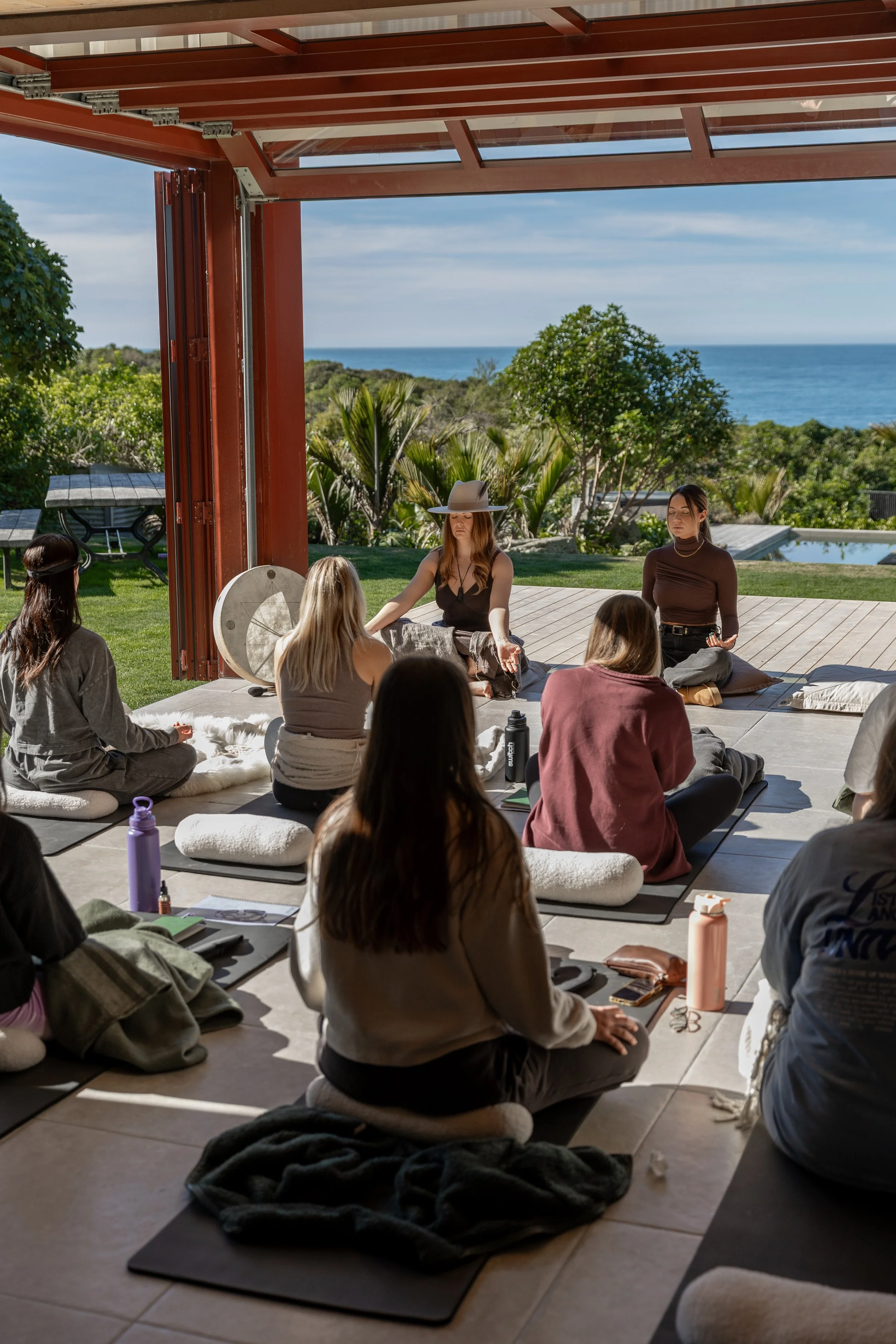 Group of people practicing meditation outdoors on a patio overlooking the ocean, sitting on mats with their eyes closed.
