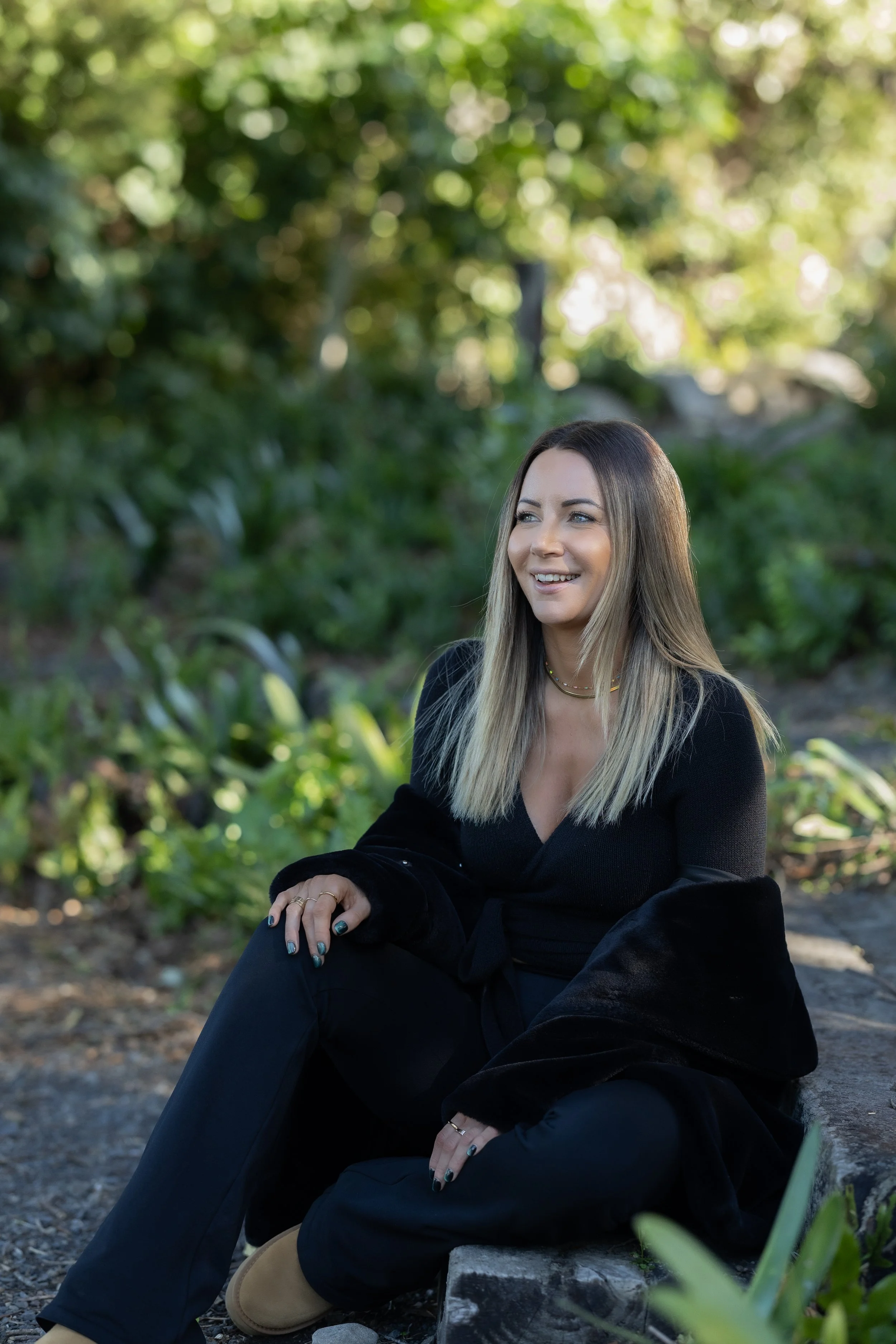 A woman with long blonde hair sitting outdoors on a wooded area with green trees and plants in the background, wearing a black outfit and smiling.