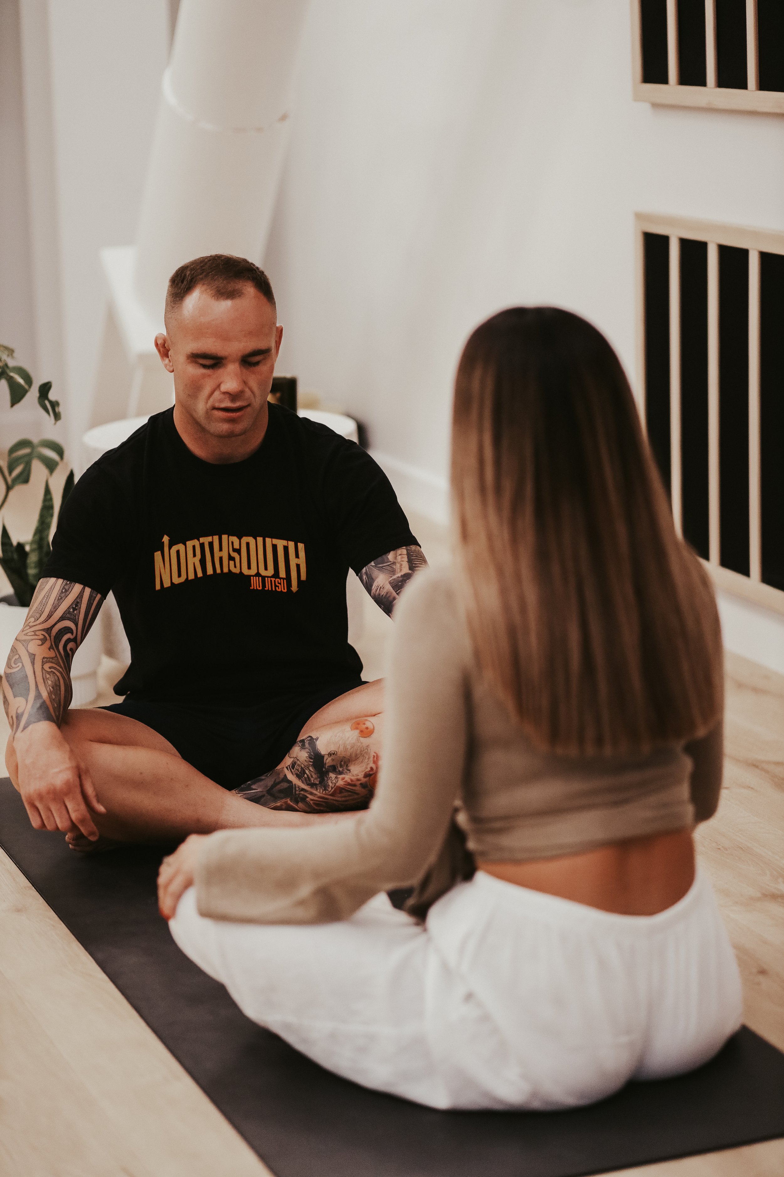 A man and woman sitting cross-legged on a yoga mat indoors, facing each other, engaging in a conversation or meditation, with a plant and window in the background.