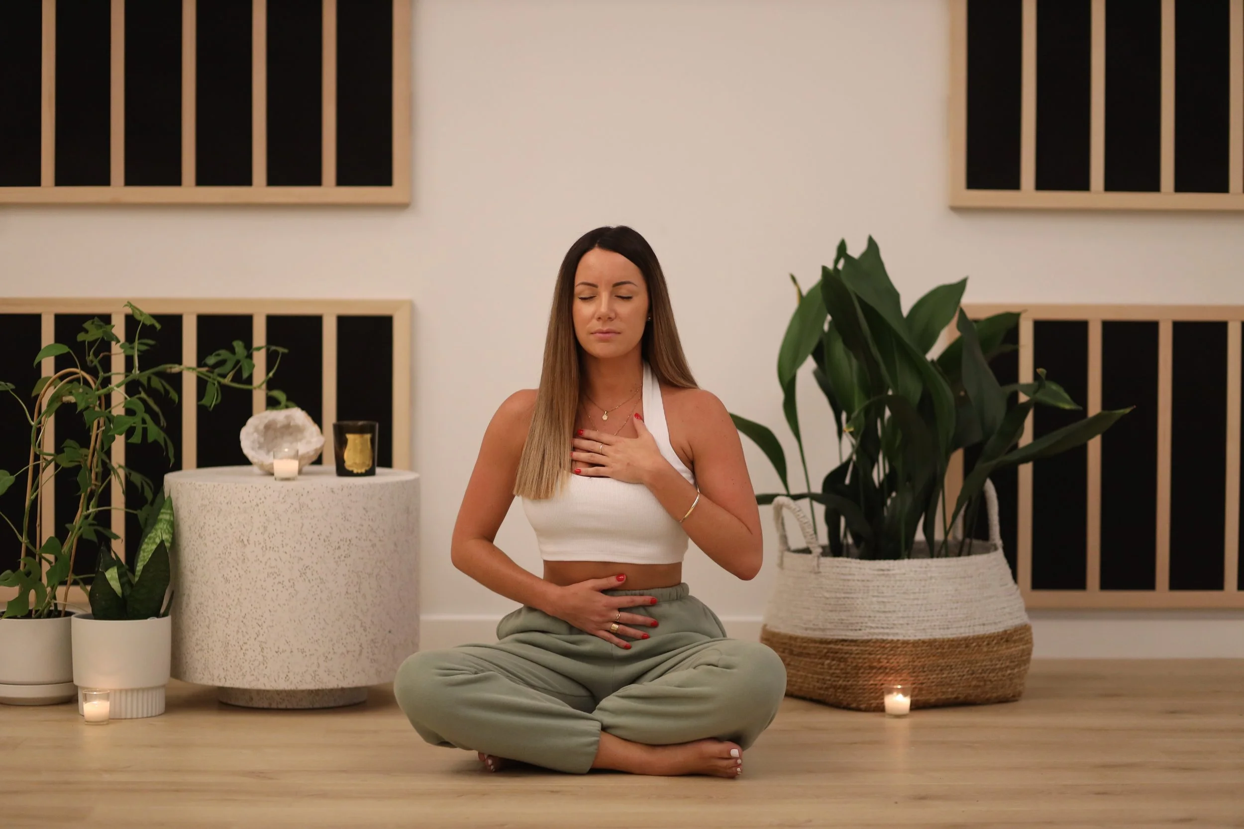 A woman practicing yoga or meditation indoors, sitting cross-legged on the floor with her eyes closed, hands on her chest and abdomen, surrounded by plants, candles, and minimalist decor.