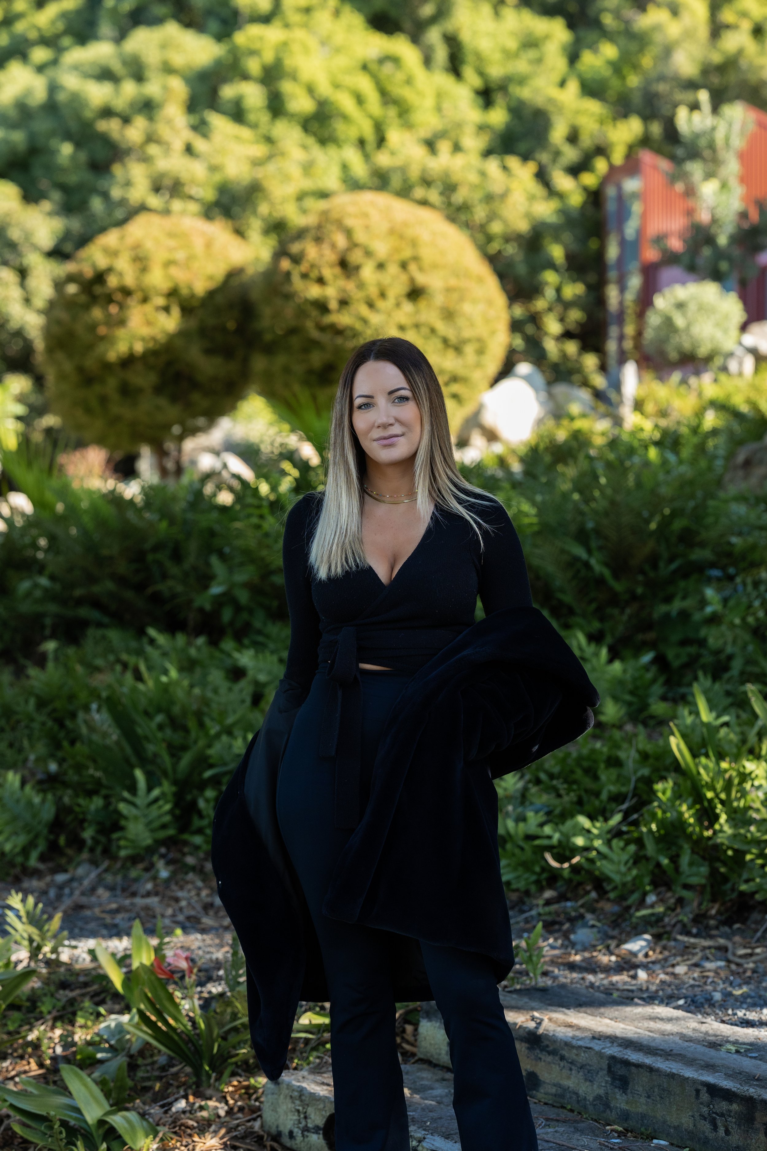 A woman with long blonde hair, wearing a black outfit, standing outdoors amidst green plants and trees.