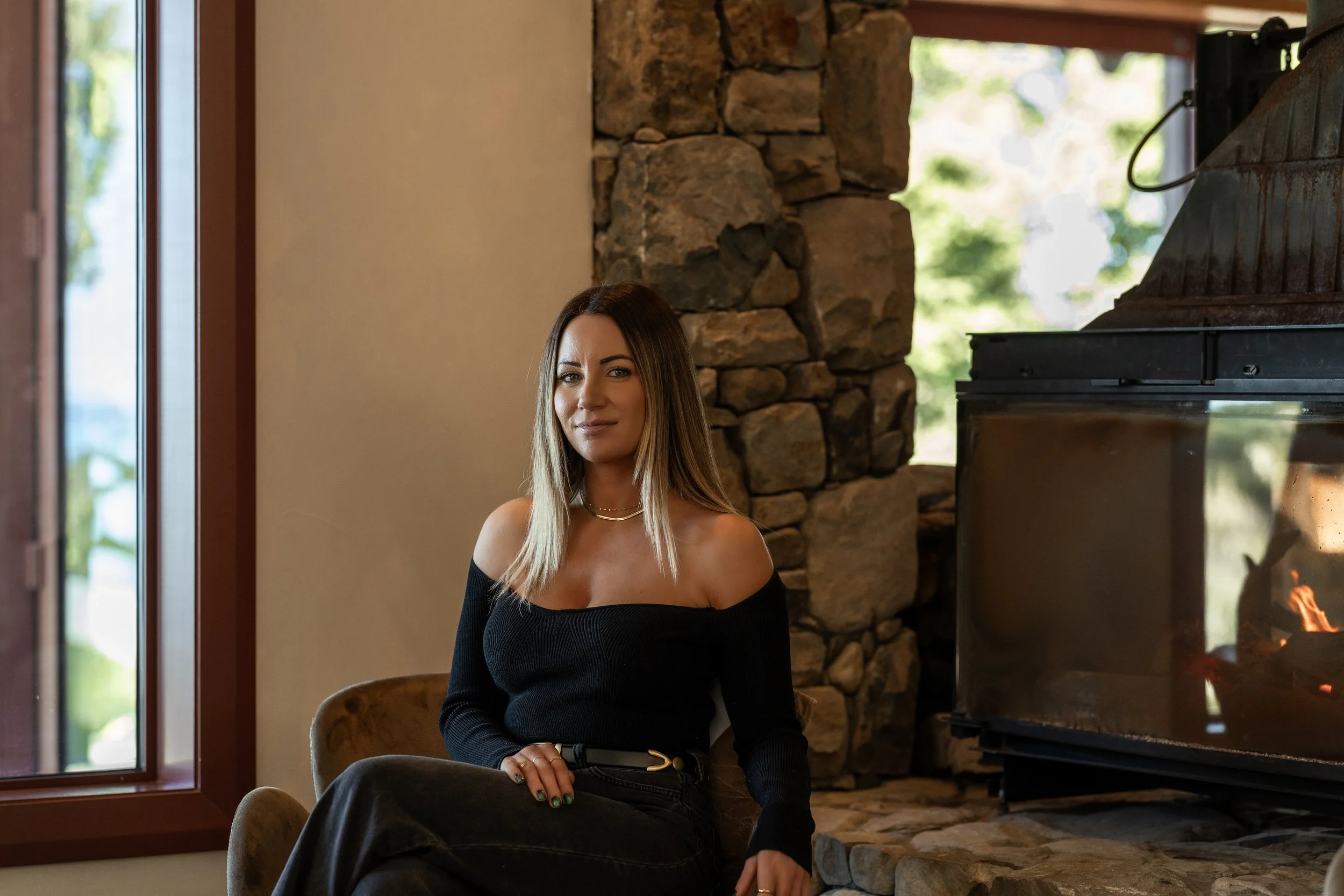 A woman with shoulder-length hair and a black off-the-shoulder top sitting near a fireplace in a cozy room with natural light.