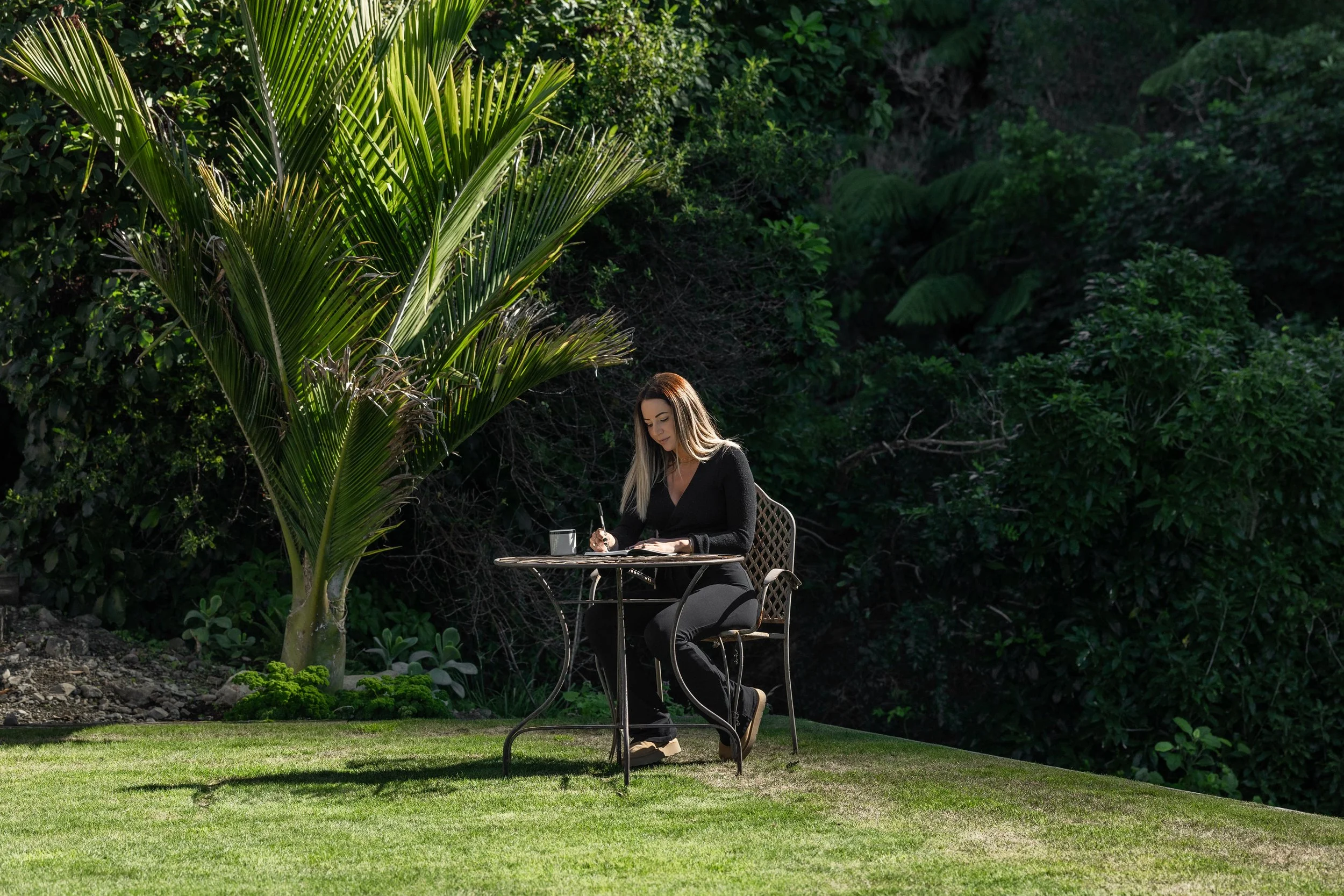 Woman sitting at an outdoor table, writing in a notebook with a cup of coffee nearby, surrounded by lush green plants and trees.