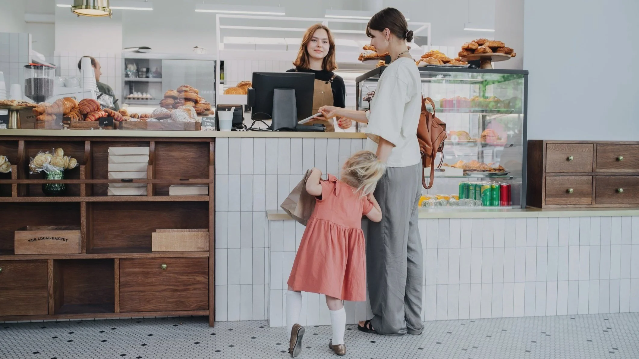 A woman with a brown backpack and a young girl with blond hair in an orange dress are at a bakery counter with baked goods, while a cashier assists them.