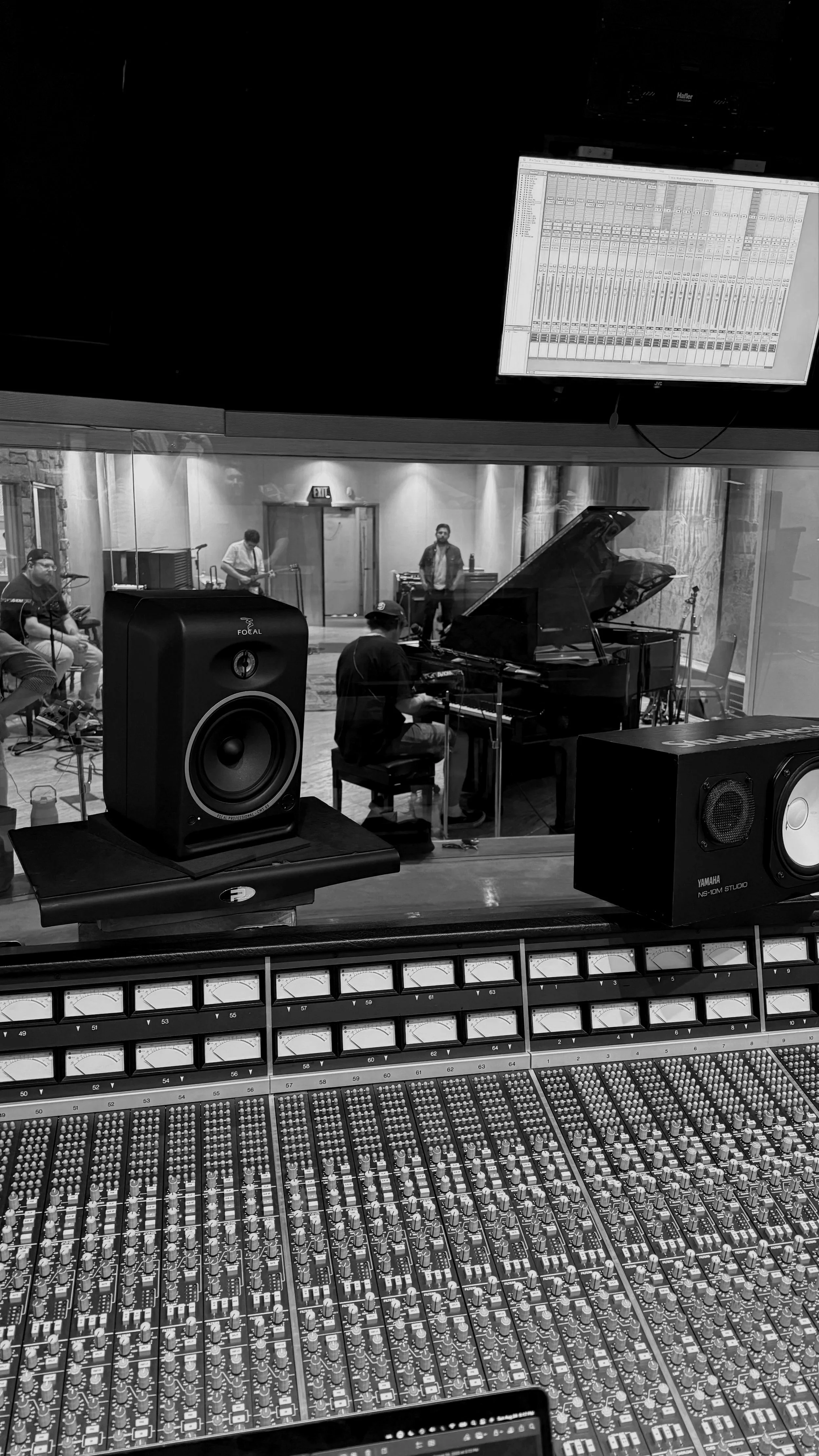 In a recording studio, musicians perform live, including a pianist at a grand piano, with a mix console in the foreground and a large screen displaying audio levels above.