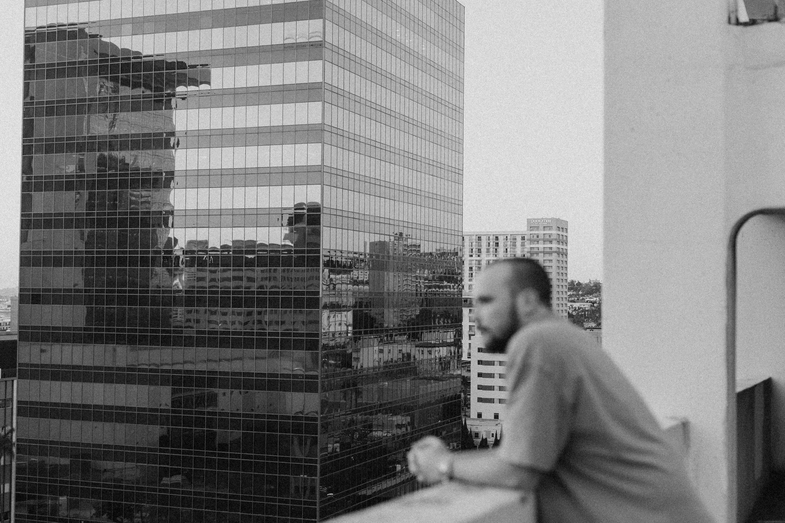 Black and white photograph of a modern glass building with reflections, with two men in the foreground, one walking and the other blurred, and distant high-rise buildings.