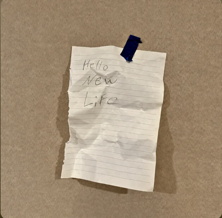 A crumpled piece of lined notebook paper taped to a beige wall with blue tape, with the handwritten words "Hello New Life".