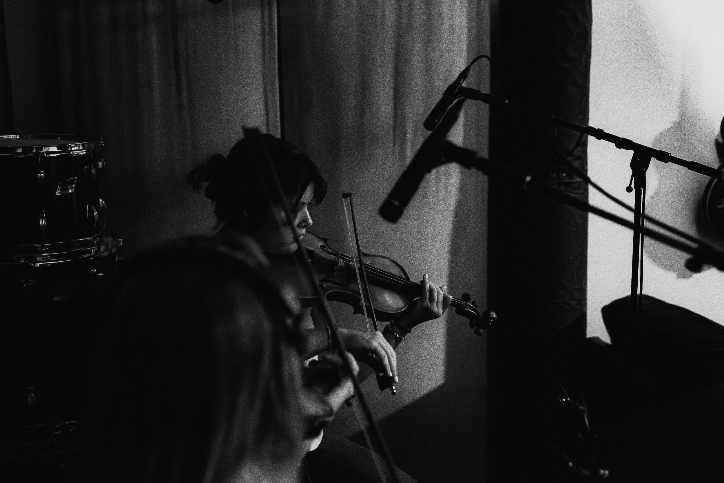 Black and white image of a woman playing the violin, with a tambourine visible to her left and microphone stands nearby. She appears to be performing on stage or in a recording studio.