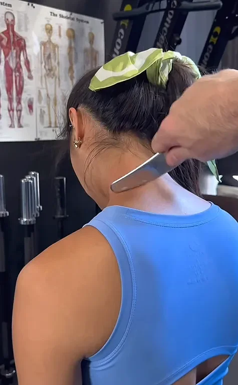 A sports massage therapist in Liverpool uses a muscle scraper tool on the neck of a woman wearing a blue top and green hair scrunchie.