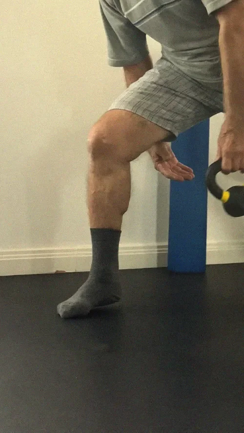 An older man wearing plaid shorts performs a weighted calf raise using a kettlebell in Liverpool exercise room.