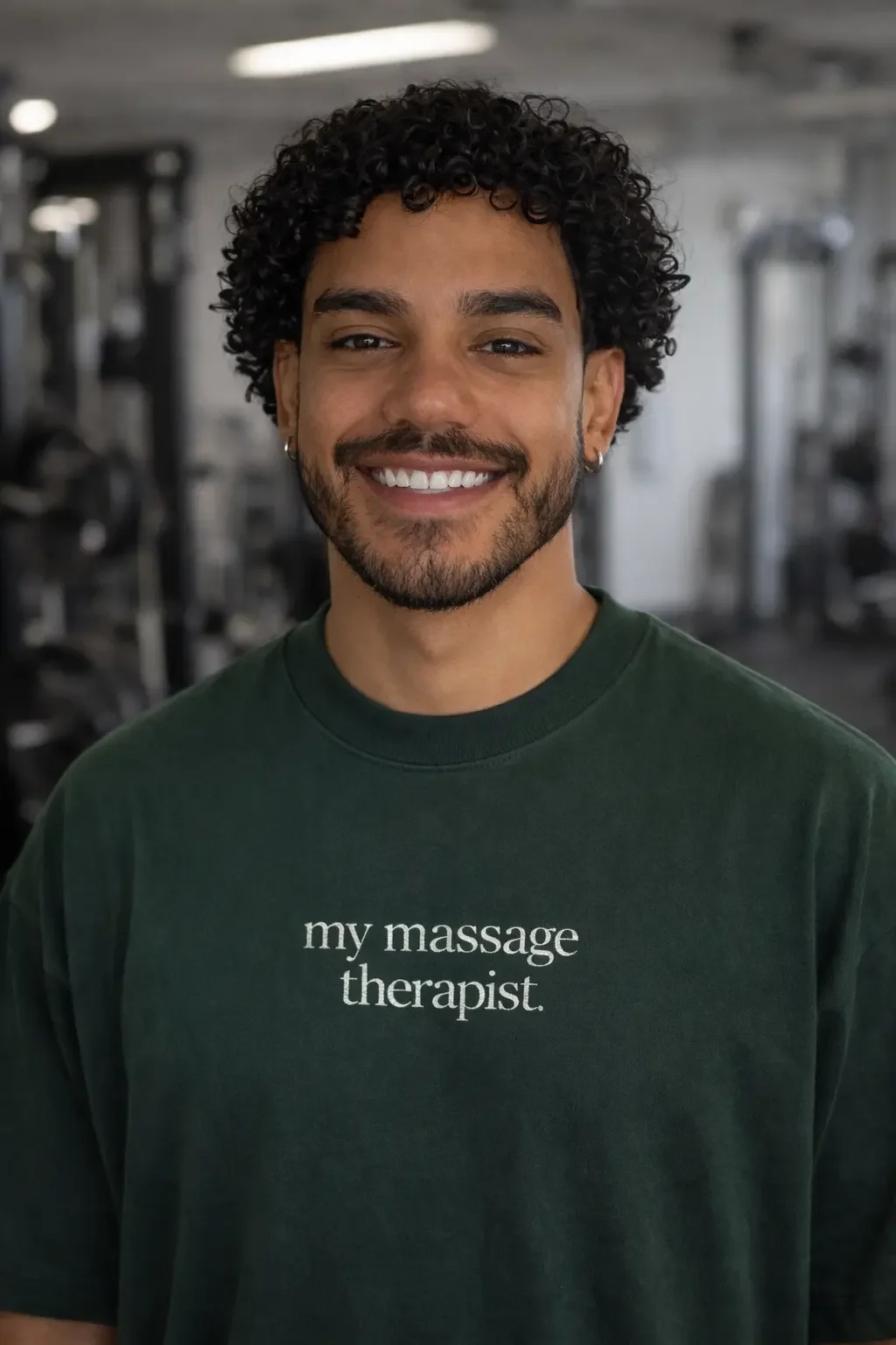 Sports massage therapist with curly black hair, beard, and earrings wearing a dark green t-shirt that says 'my massage therapist,' standing in a gym or fitness center located in Liverpool.