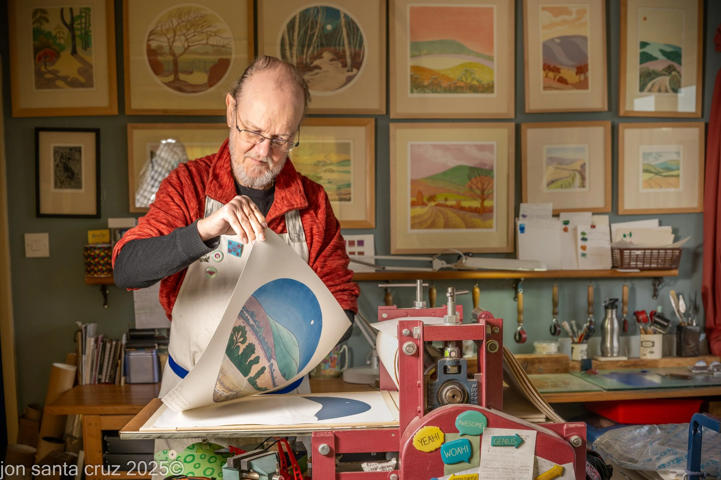 Ian O'Halloran, Artist Printmaker in his studio peeling off a print after it has gone through the printing press. His framed works on wall behind.