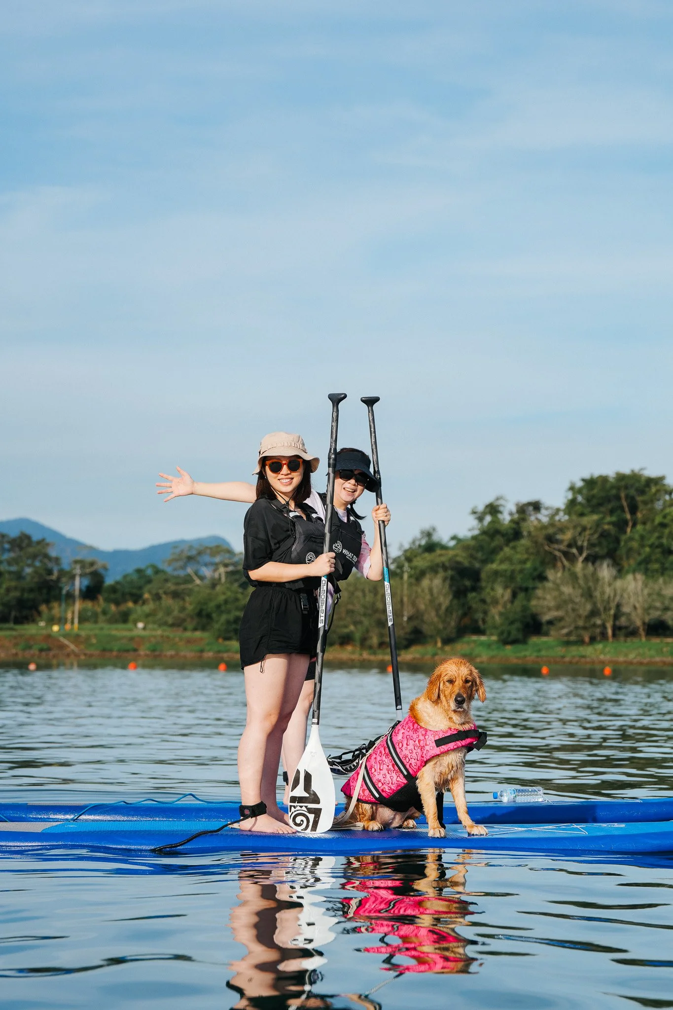 Two women stand on a paddleboard with a dog during a sunny day on a lake. The women are smiling, holding paddles, and wearing sunglasses and hats. The dog, sitting on the paddleboard, is wearing a pink life vest.