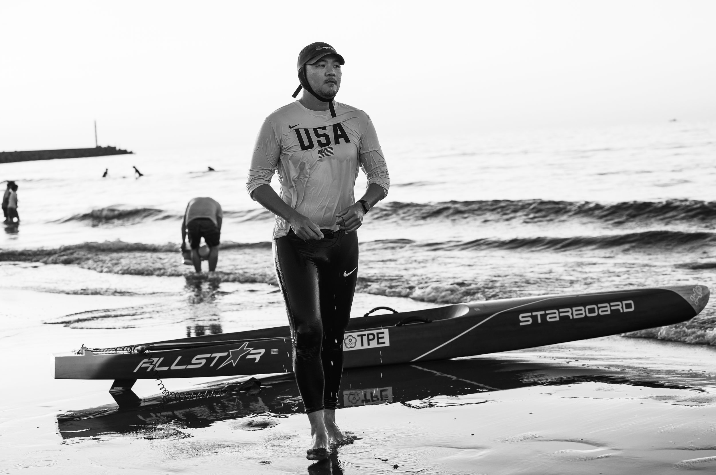 Male athlete wearing a USA jersey and helmet stands on the beach next to a paddleboard labeled 'STARBOARD' with a Taiwan flag sticker, with other people in the water in the background.