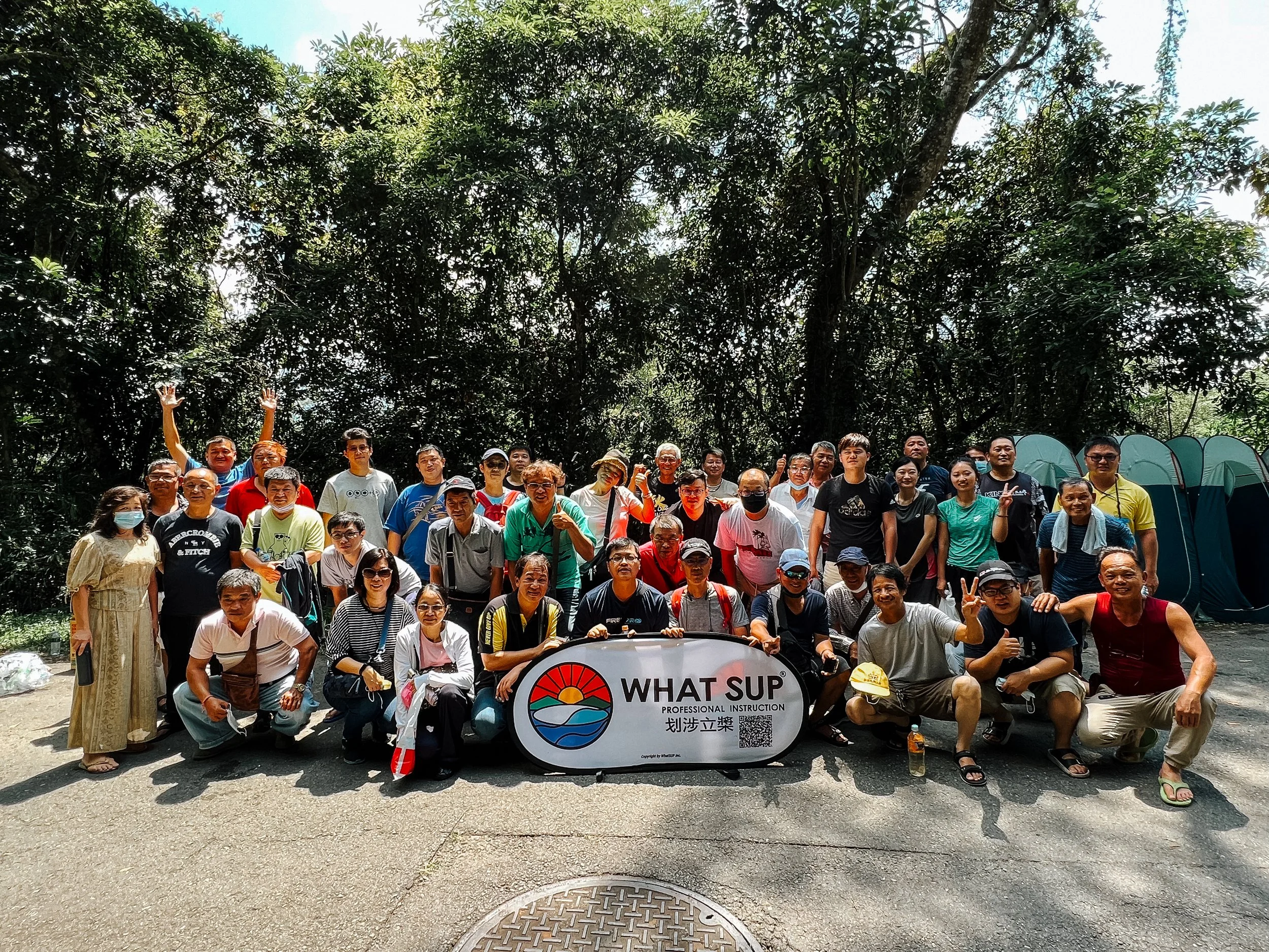 Group of people outdoors holding a banner that reads 'WHAT SUP' with a sunset and water graphic, surrounded by trees and tents in the background.