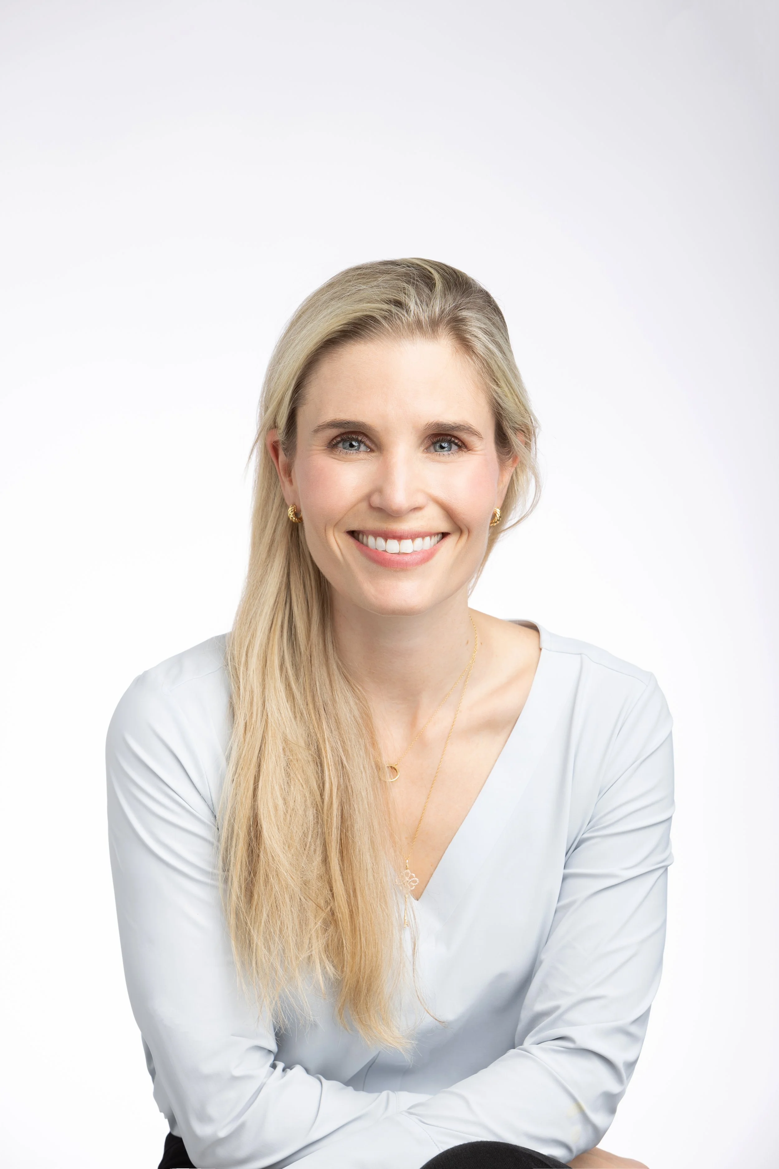 A woman with long blonde hair smiling, wearing a white blouse and gold jewelry against a plain white background.