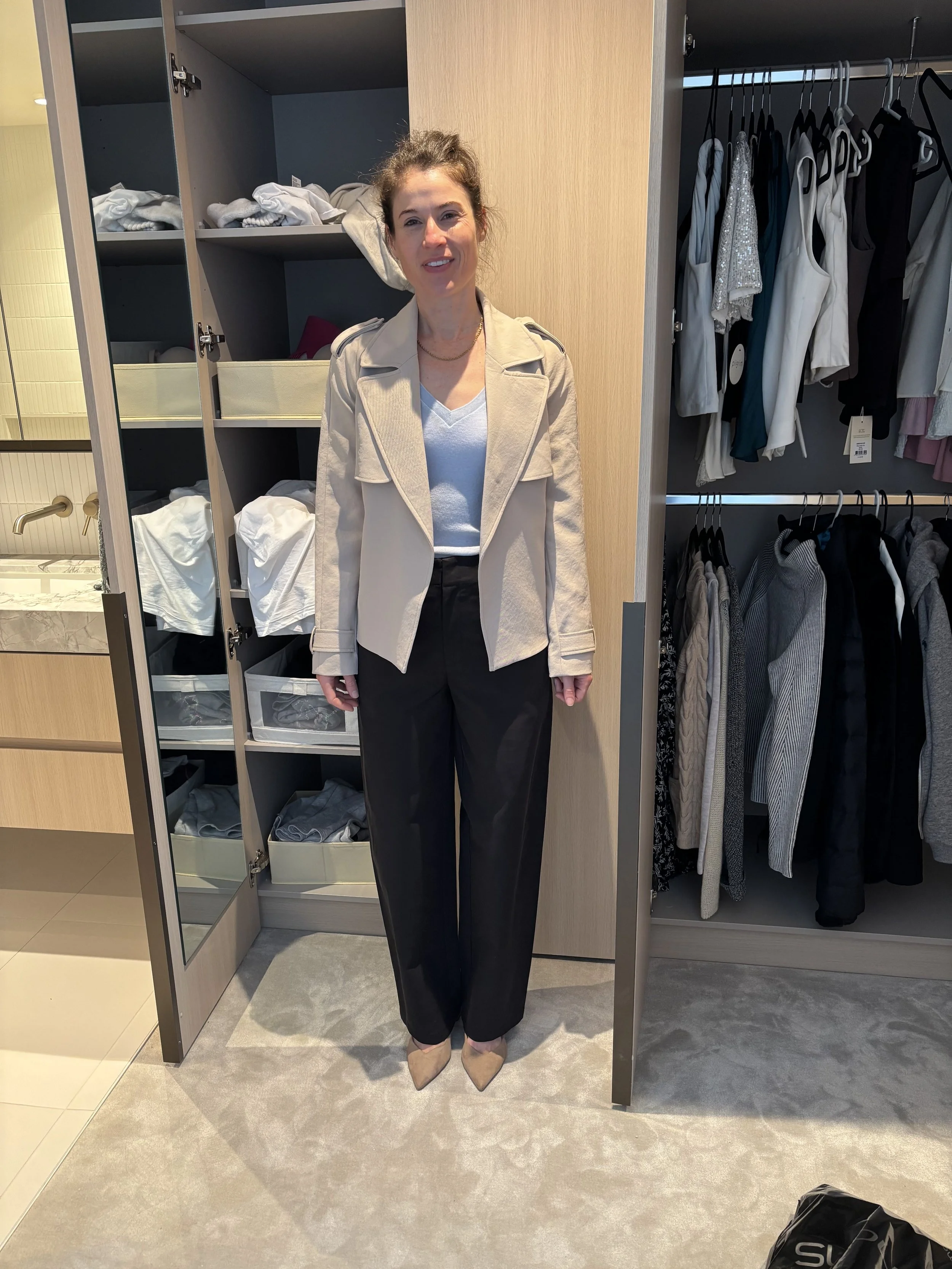 A woman standing in front of an open wardrobe with folded clothes on the left and hanging clothes on the right. The woman is wearing a beige jacket, a light blue top, black pants, and beige high heels.