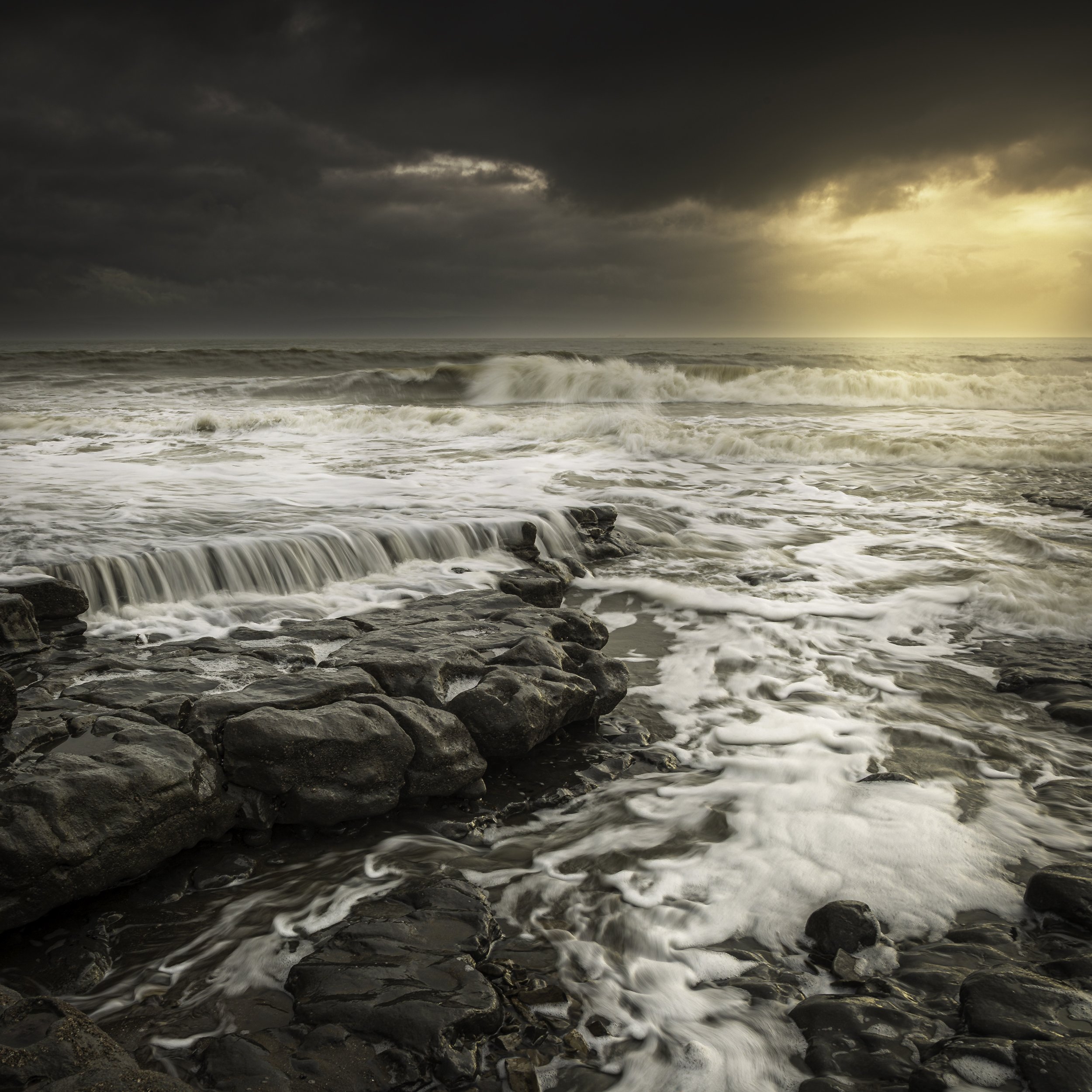 Ocean waves crashing on rocky shore with dramatic cloudy sky and sunlight breaking through. Foam and water flowing over rocks.
