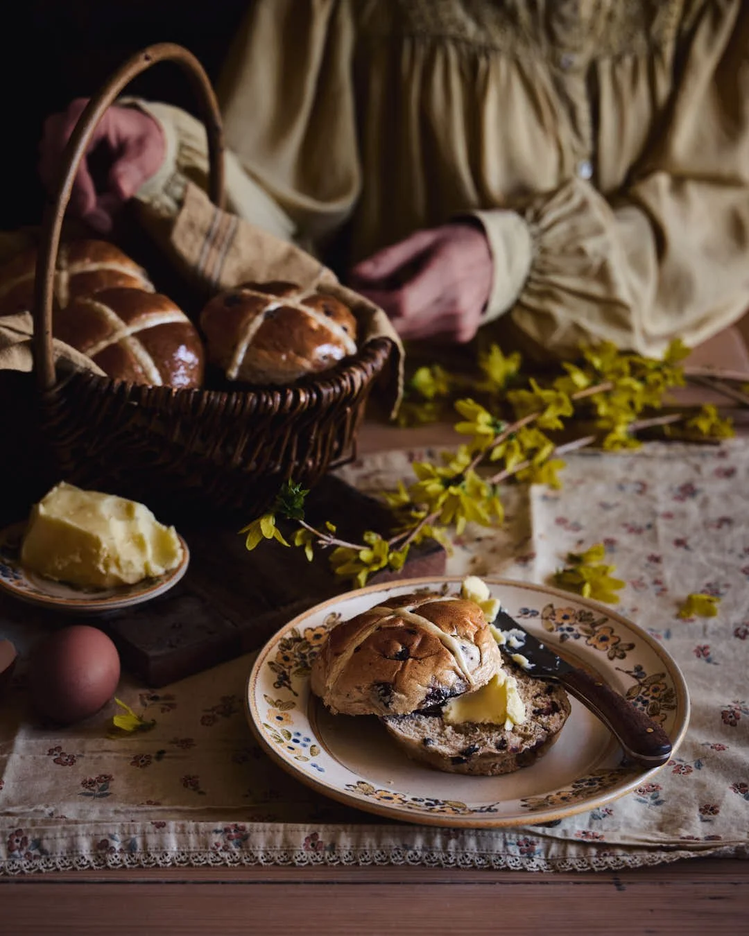 angled image of hot cross bun on plate with butter and girl in yellow blouse behind