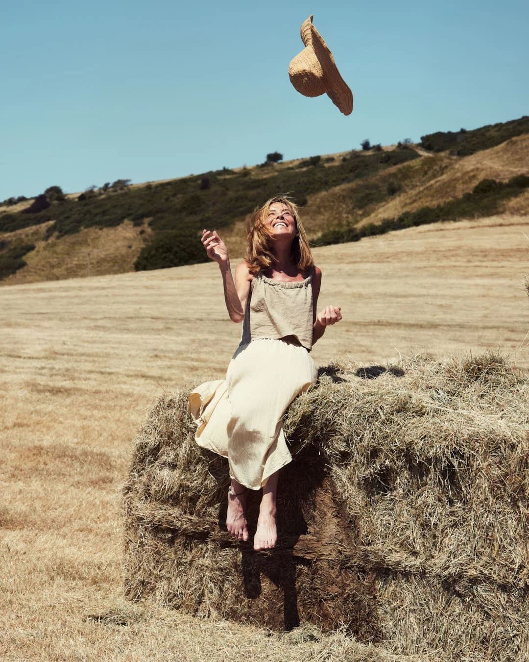 girl throwing straw hat in air on hay bale in summer sun