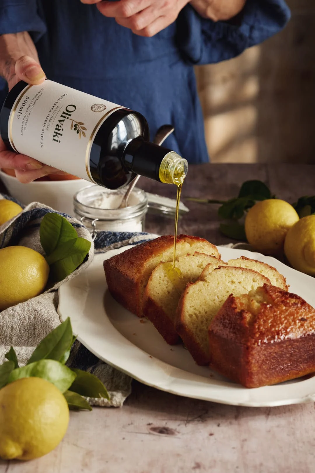 angled  image of olive oil being poured onlemon loaf cake surrounded by lemons on rustic wood surface