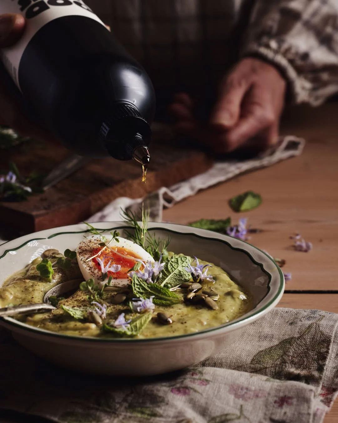 angled image of olive oil being drizzled onto green soup in rustic farmhouse setting
