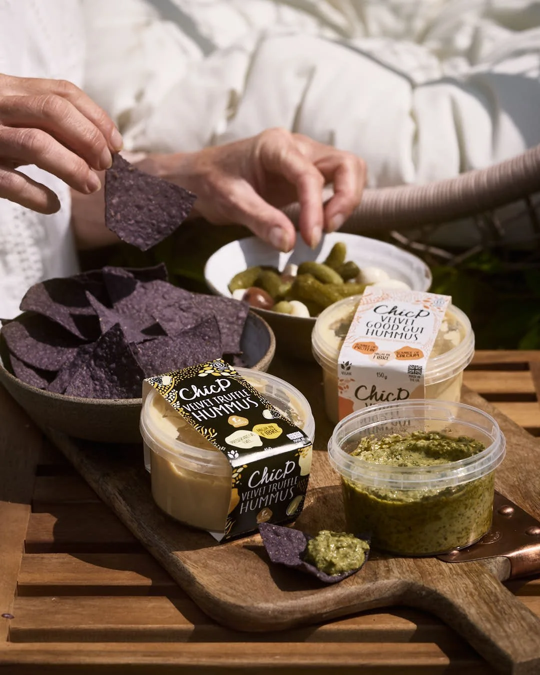 chickpea hummus iun plastic pots in summer scene with purple tortillas and hand reaching for pickles outdoors