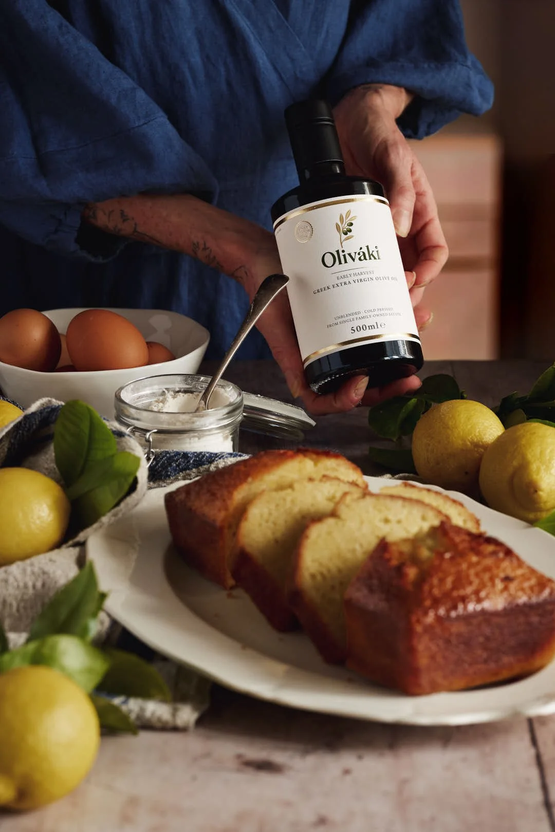 angled  image of girl holding olive oil bottle by lemon loaf cake surrounded by lemons on rustic wood surface
