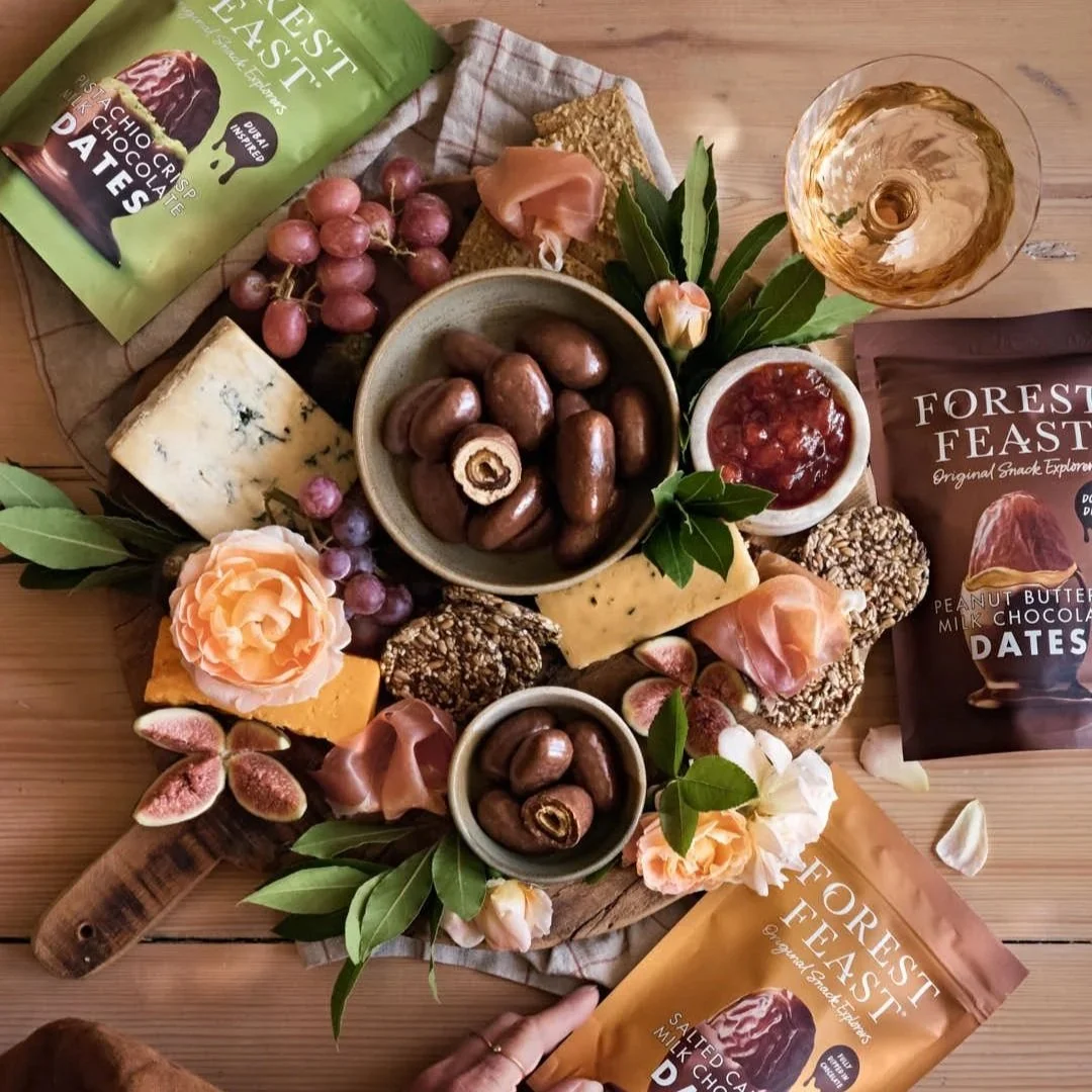 flatlay image of serving board with dates and flowers and artisan cheeses with chocolate and packaging