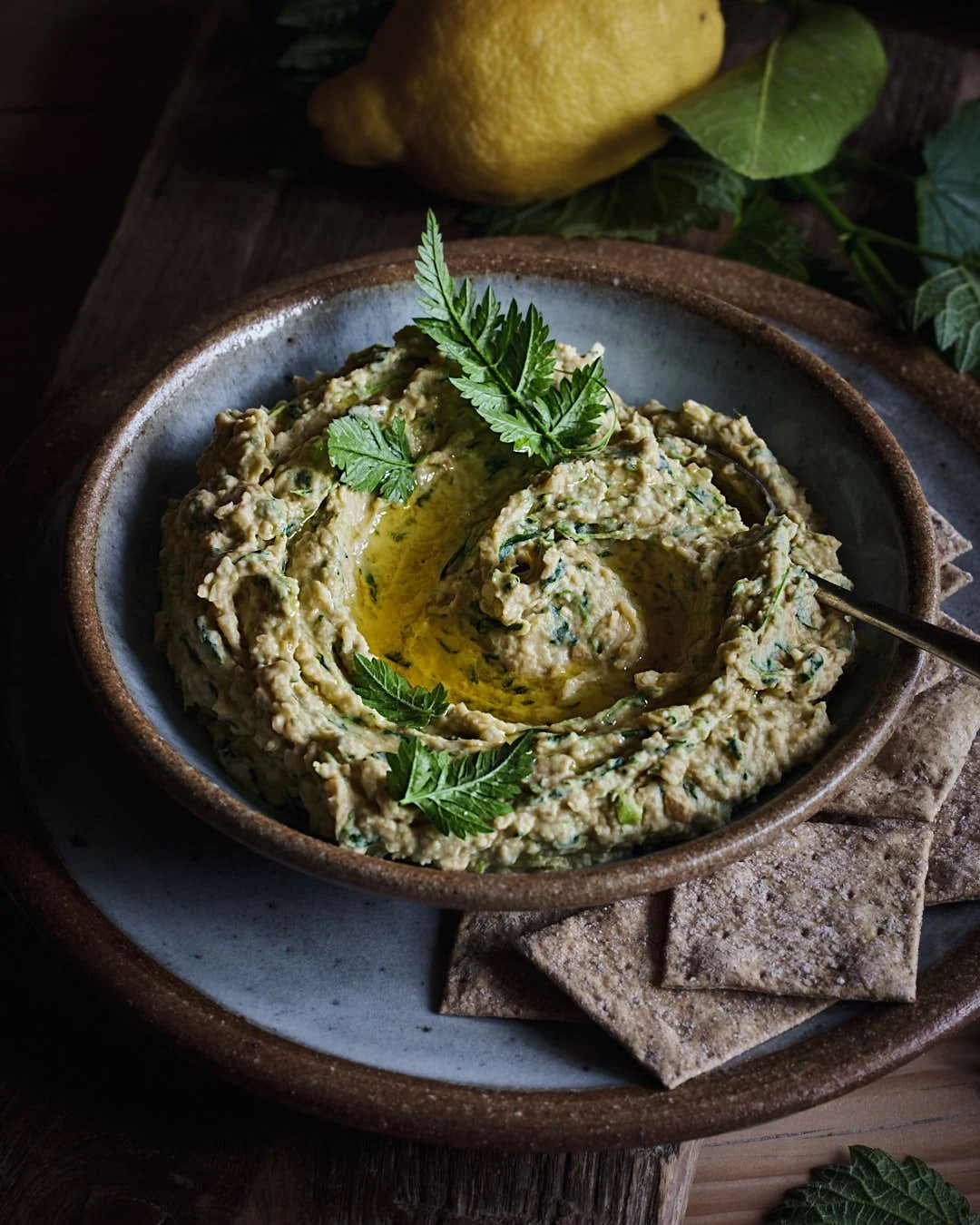 angled image of hummus in bowl to show chunky texture and blue shadows with lemons in rustic moody lit scene