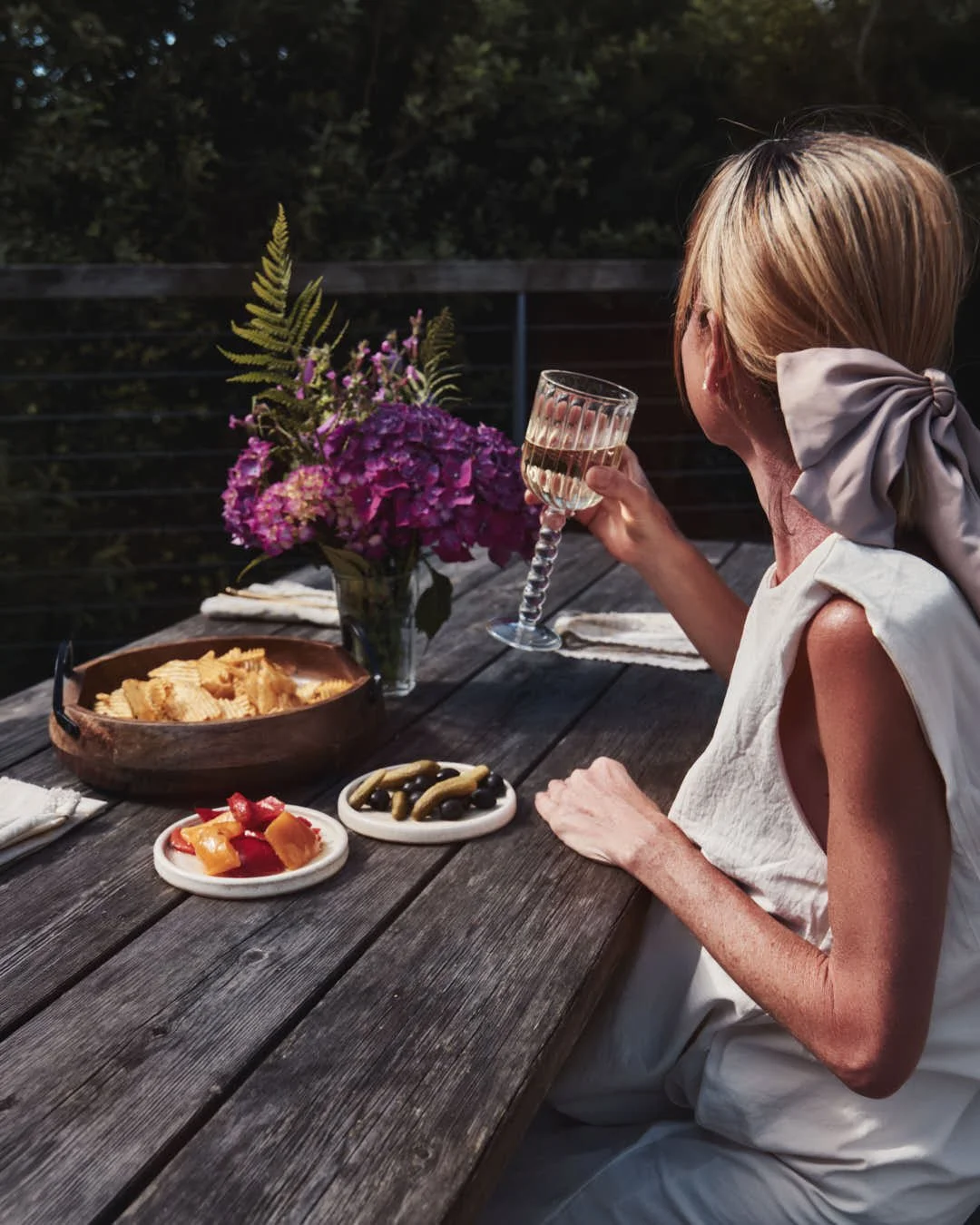 girl holding glass to sun at outside table with flowers and nibbles in sunlit scene