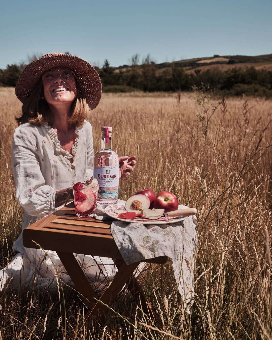 girl in sun dress in summer sun staring at sky in field with small table gin and glass with nectarines