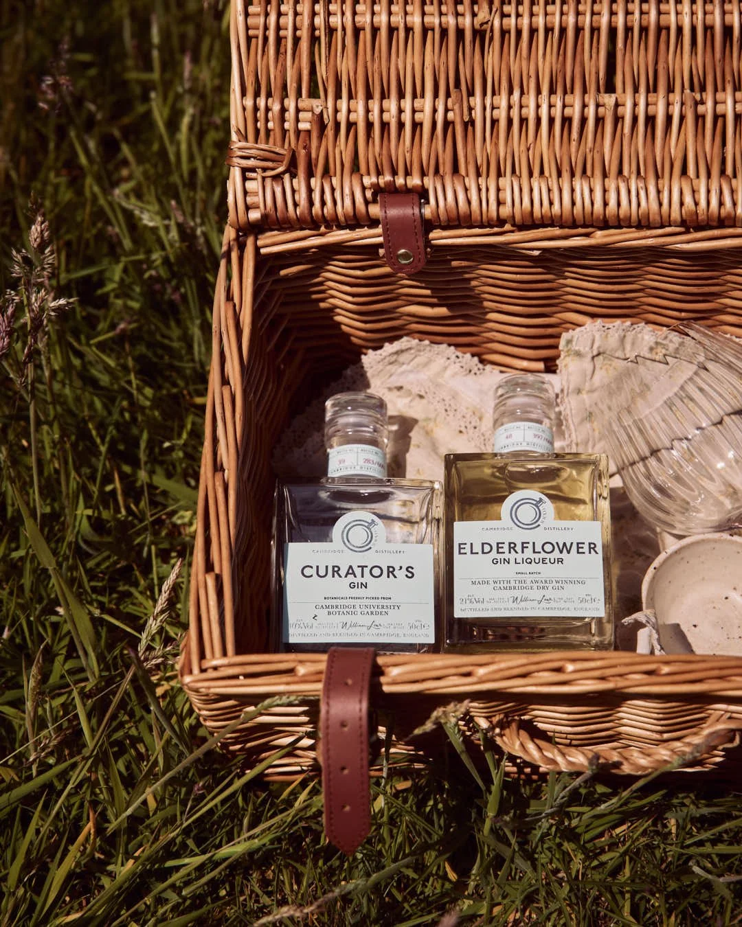 overhead image of two gin bottles in open wicker picnic hamper in summer sunshine