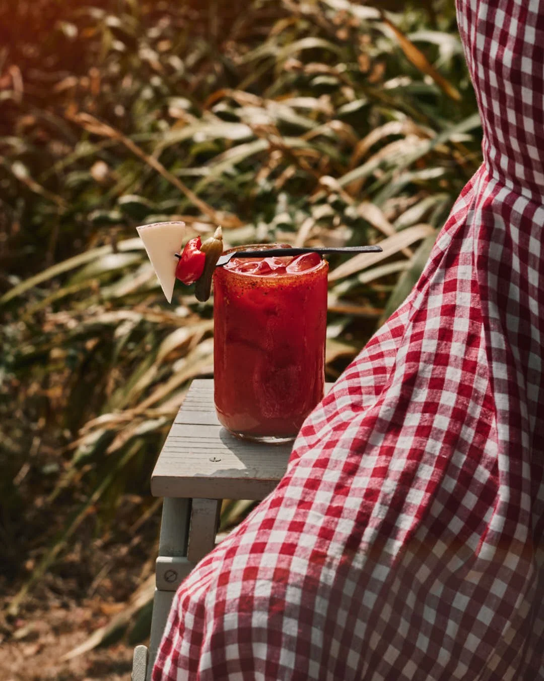 angled image of tomato drink in bright summer sun with gigham dress behind and mini peppers on plate